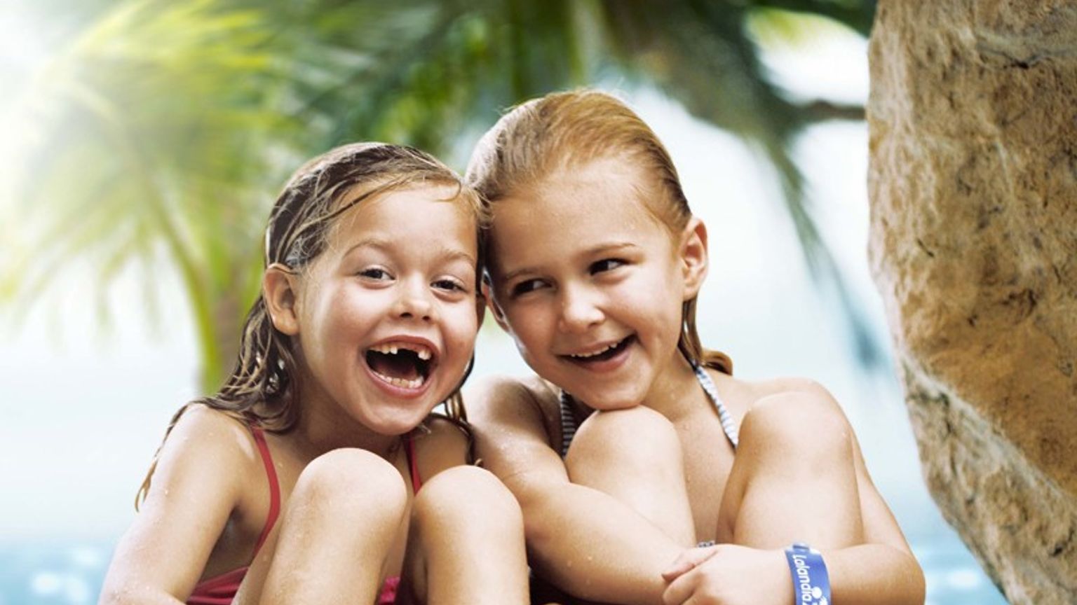 To glade unge piger med vådt hår og blå armbånd sidder på klipper i en tropisk grotte med vand og palmer bag dem. Lalandia Rødby