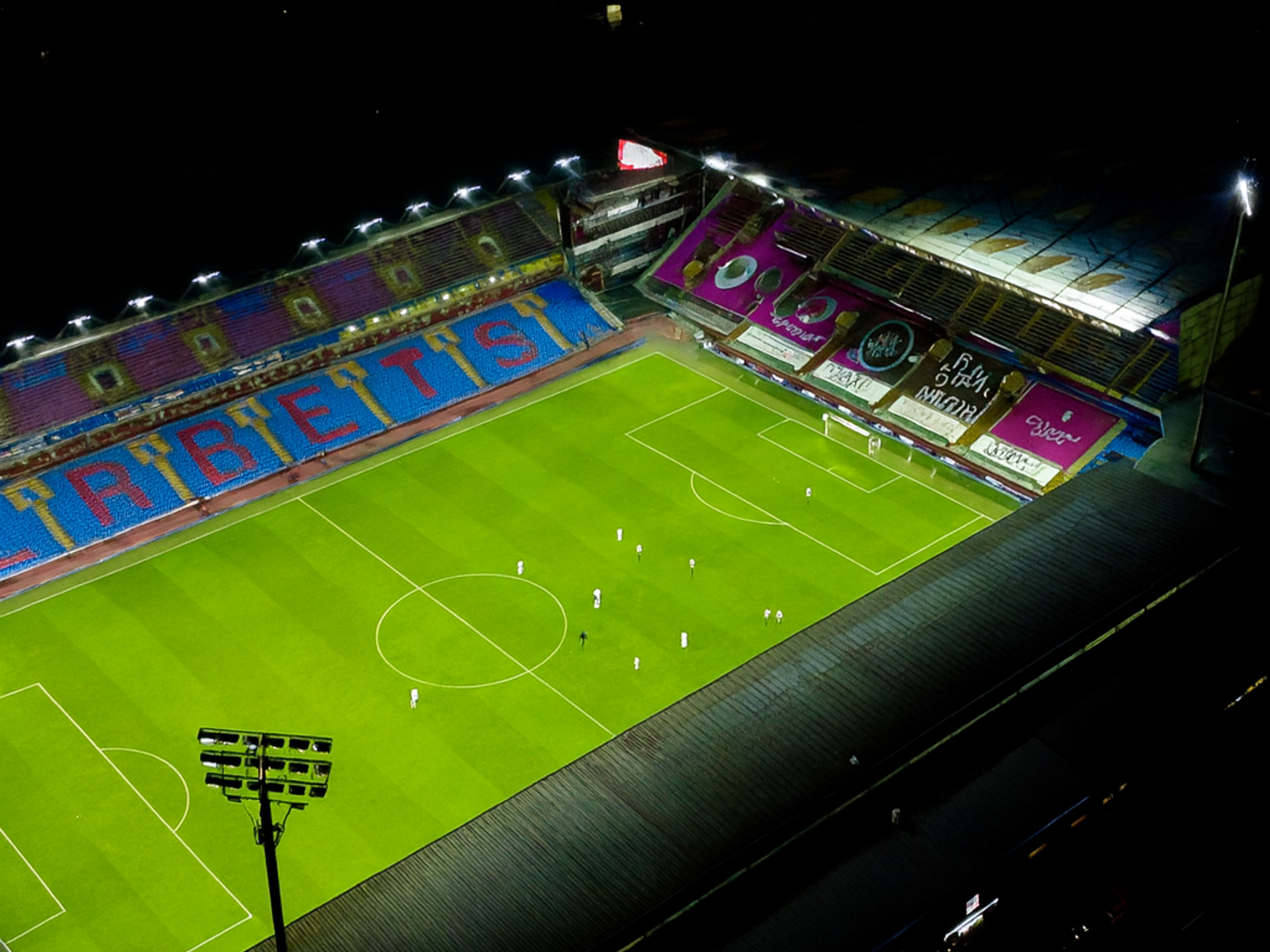 Vue aérienne d'un stade de football brillamment éclairé la nuit avec des joueurs sur le terrain vert, sous un ciel sombre avec des étoiles et des lumières flottantes colorées.