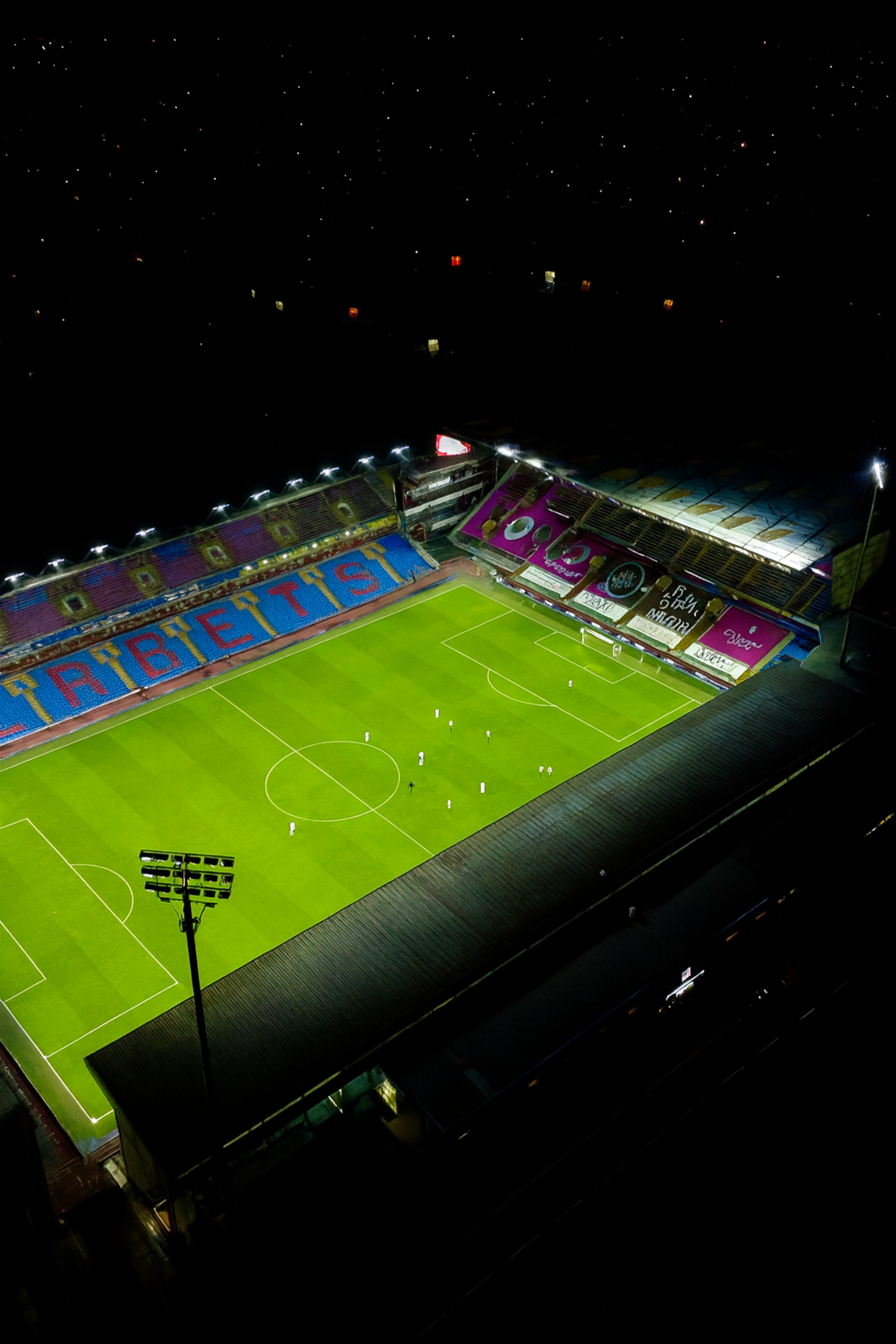Vue aérienne d'un stade de football brillamment éclairé la nuit avec des joueurs sur le terrain vert, sous un ciel sombre avec des étoiles et des lumières flottantes colorées.