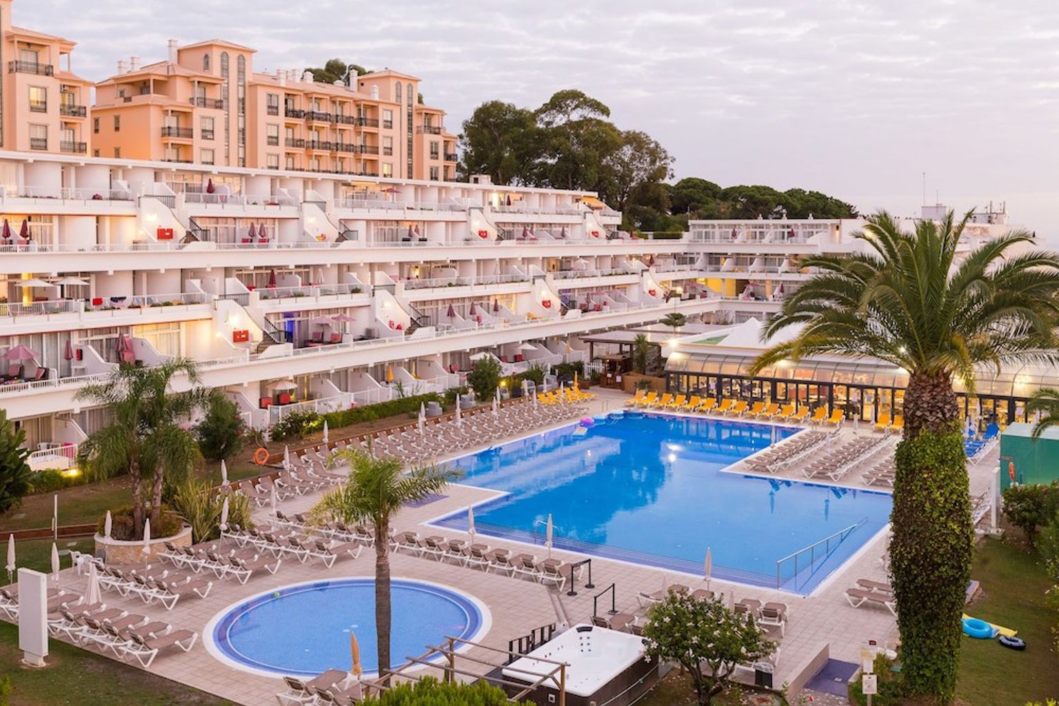 Stora terrasserade resortbyggnader vetter mot flera pooler och rader av solstolar, med palmer under en molnig himmel.