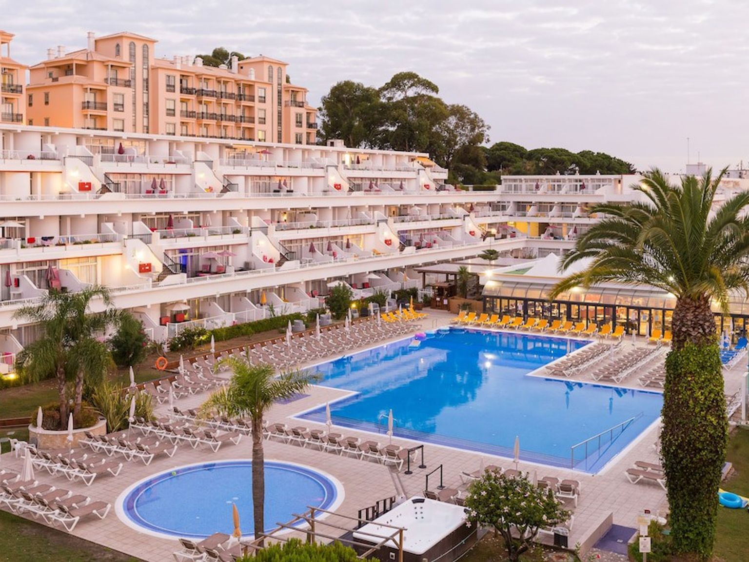 Stora terrasserade resortbyggnader vetter mot flera pooler och rader av solstolar, med palmer under en molnig himmel.