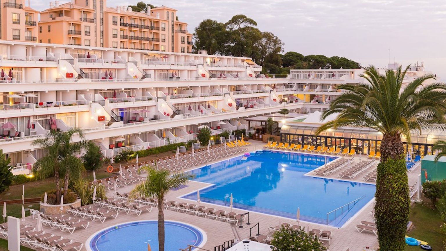 Stora terrasserade resortbyggnader vetter mot flera pooler och rader av solstolar, med palmer under en molnig himmel.