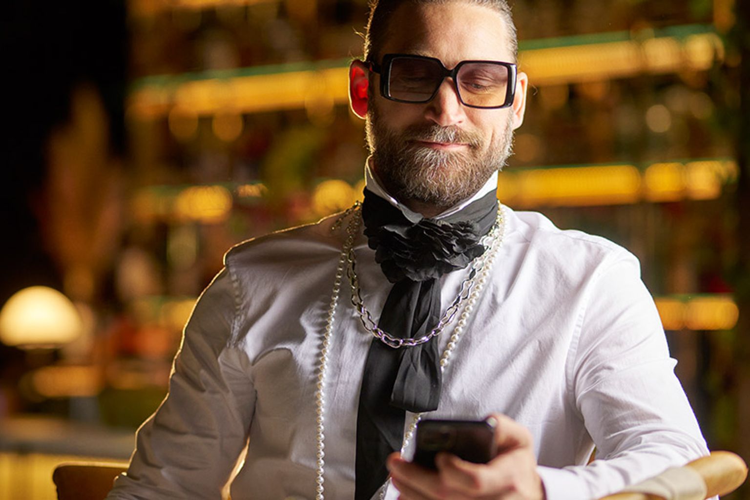 en mann med skjegg og briller sitter ved et bord og ser på mobiltelefonen sin