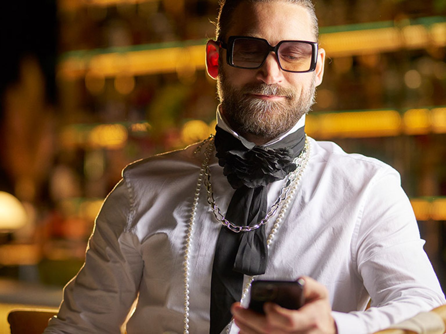 en mann med skjegg og briller sitter ved et bord og ser på mobiltelefonen sin