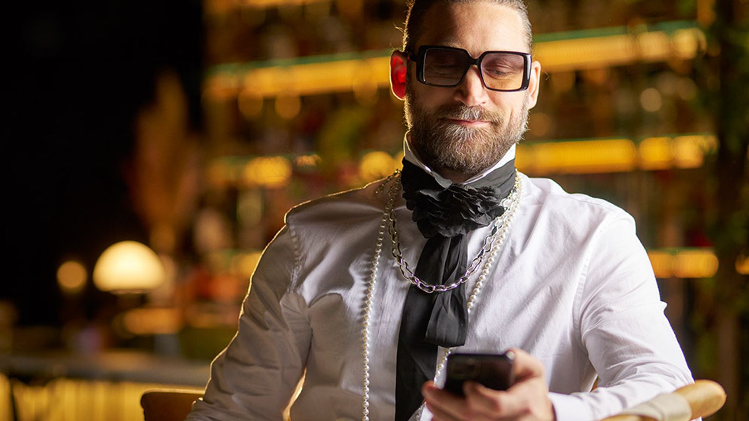 en mann med skjegg og briller sitter ved et bord og ser på mobiltelefonen sin