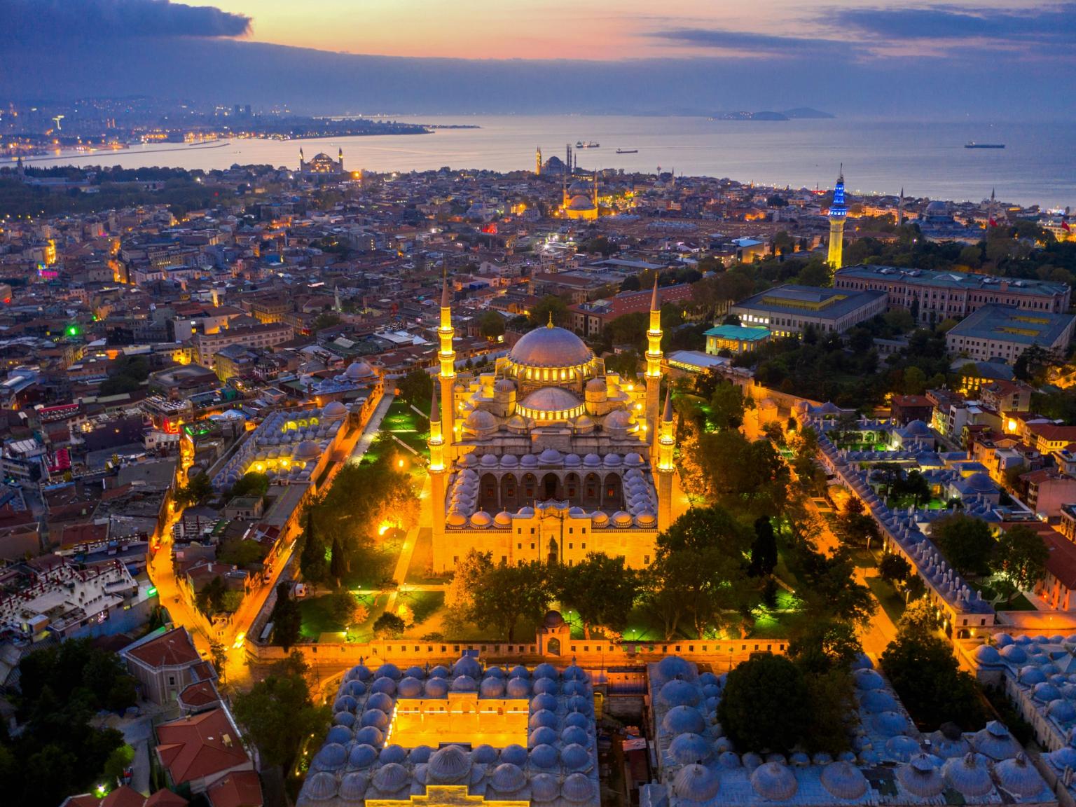 Luftfoto af den oplyste Süleymaniye-moské og Istanbuls bysilhuet i skumringen, med havet i baggrunden.