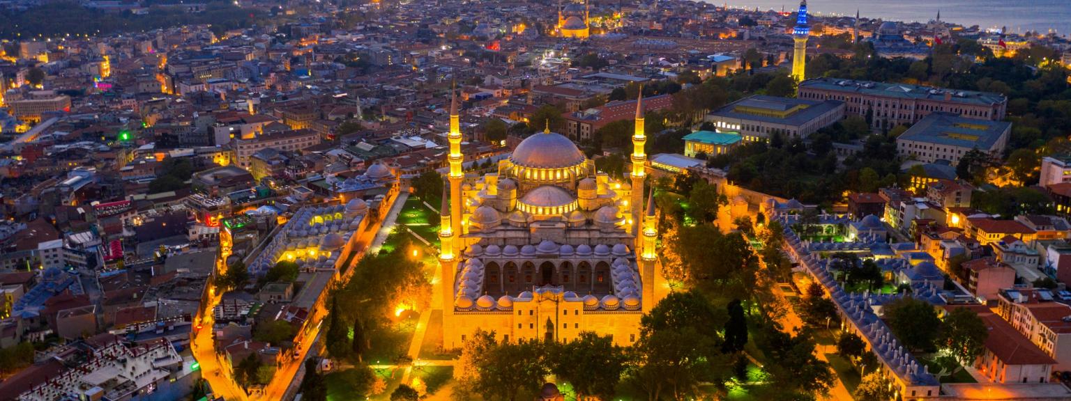 Luftfoto af den oplyste Süleymaniye-moské og Istanbuls bysilhuet i skumringen, med havet i baggrunden.