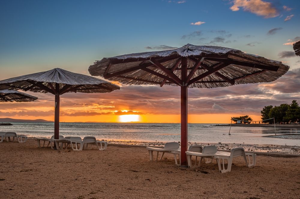 Solnedgang over en rolig strand med stråtageparaplyer og tomme liggestole. Zaton, Kroatien
