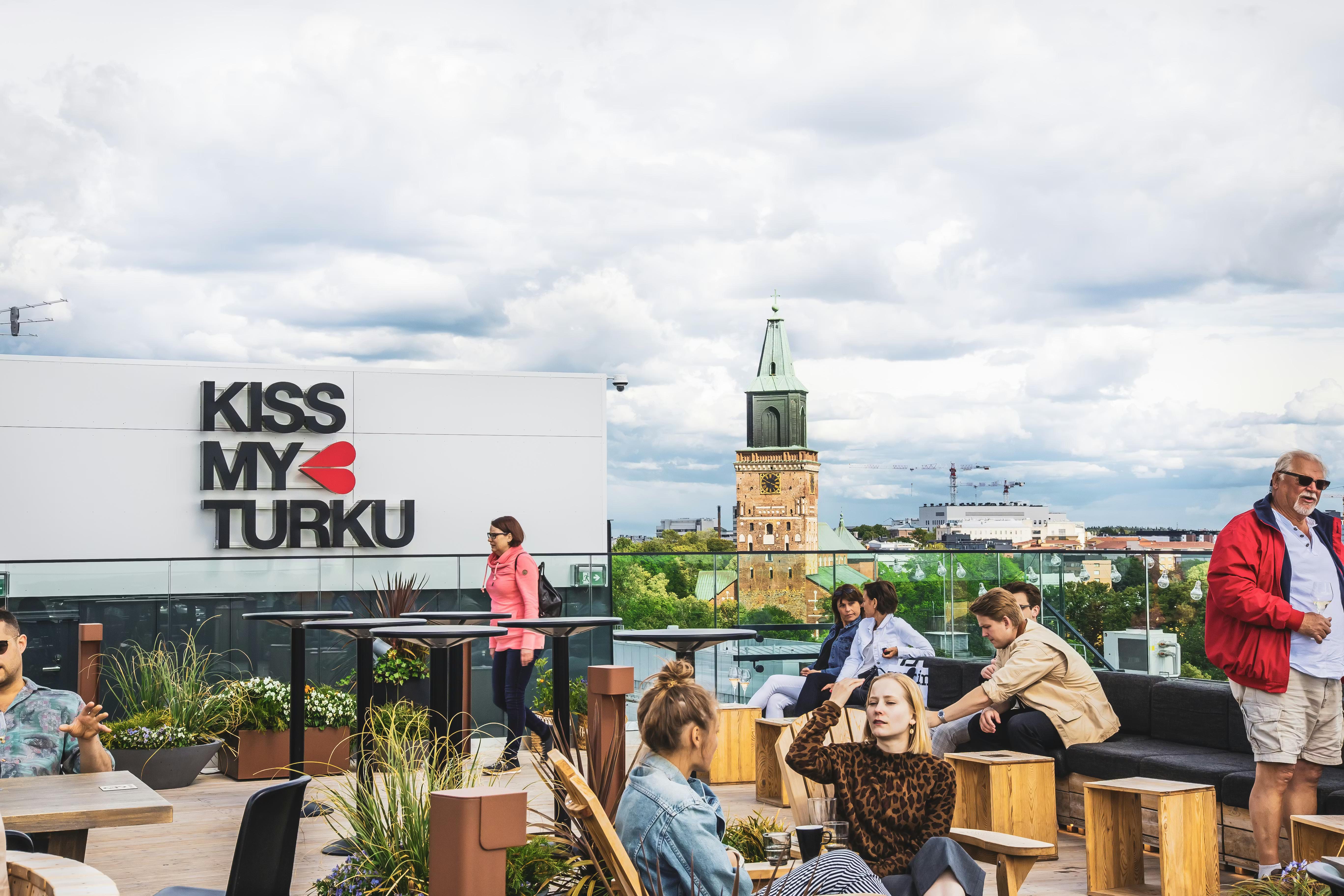 Folk slapper af på en tagterrasse med et skilt med teksten "Kiss My Turku" og udsigt til et historisk katedraltårn.