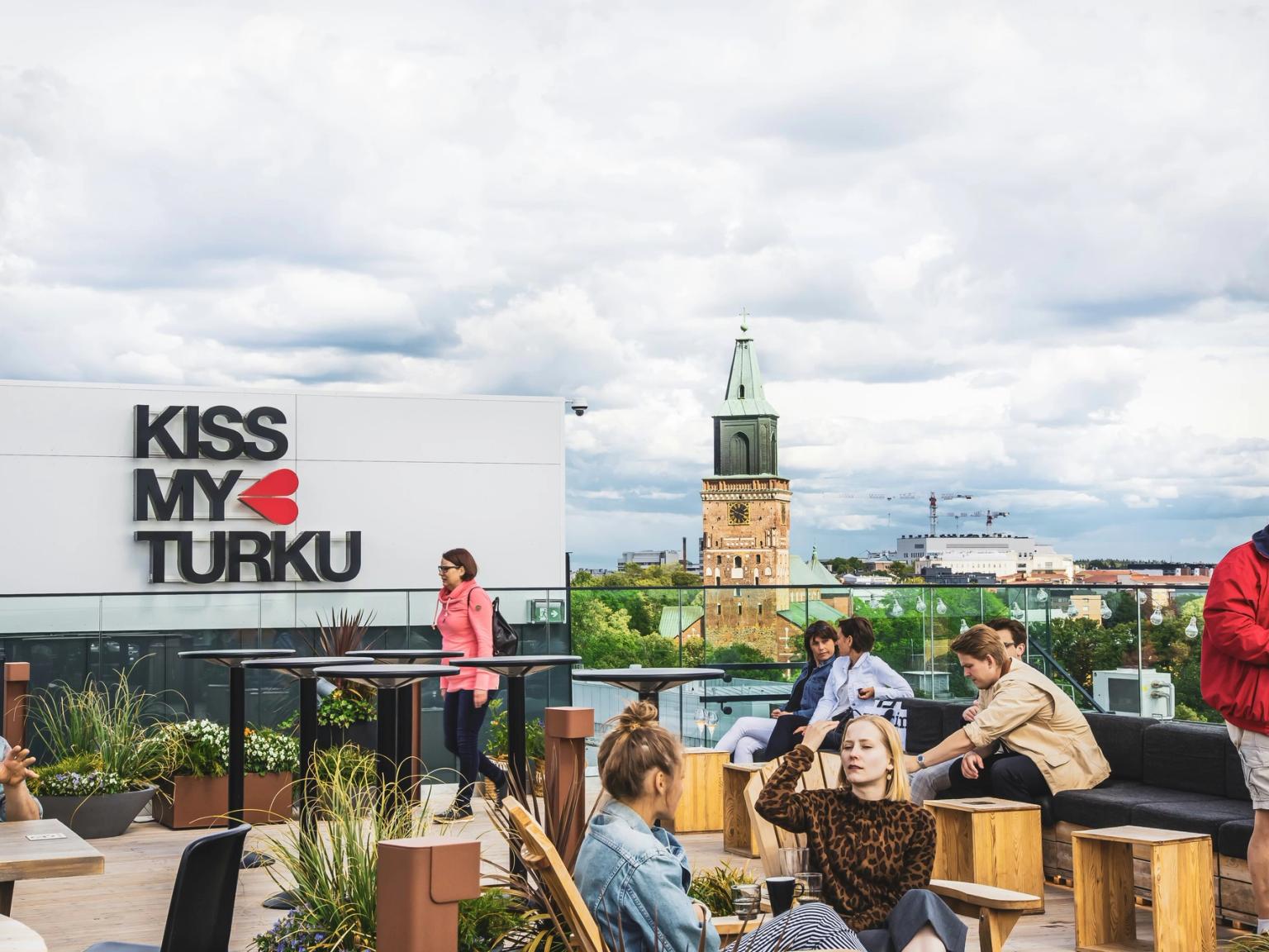 People relaxing on a rooftop terrace with a "Kiss My Turku" sign and a view of a historic cathedral tower.
