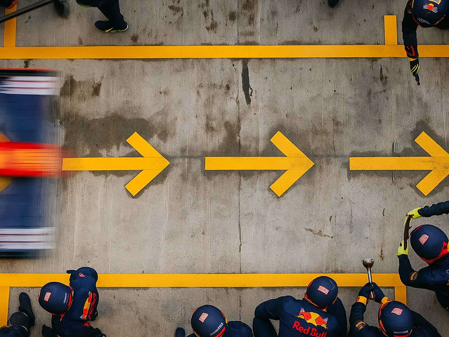 Draufsicht auf ein unscharfes F1-Auto, das an einer Red Bull-Boxencrew auf einer Betonstrecke mit gelben Richtungspfeilen vorbeirast.