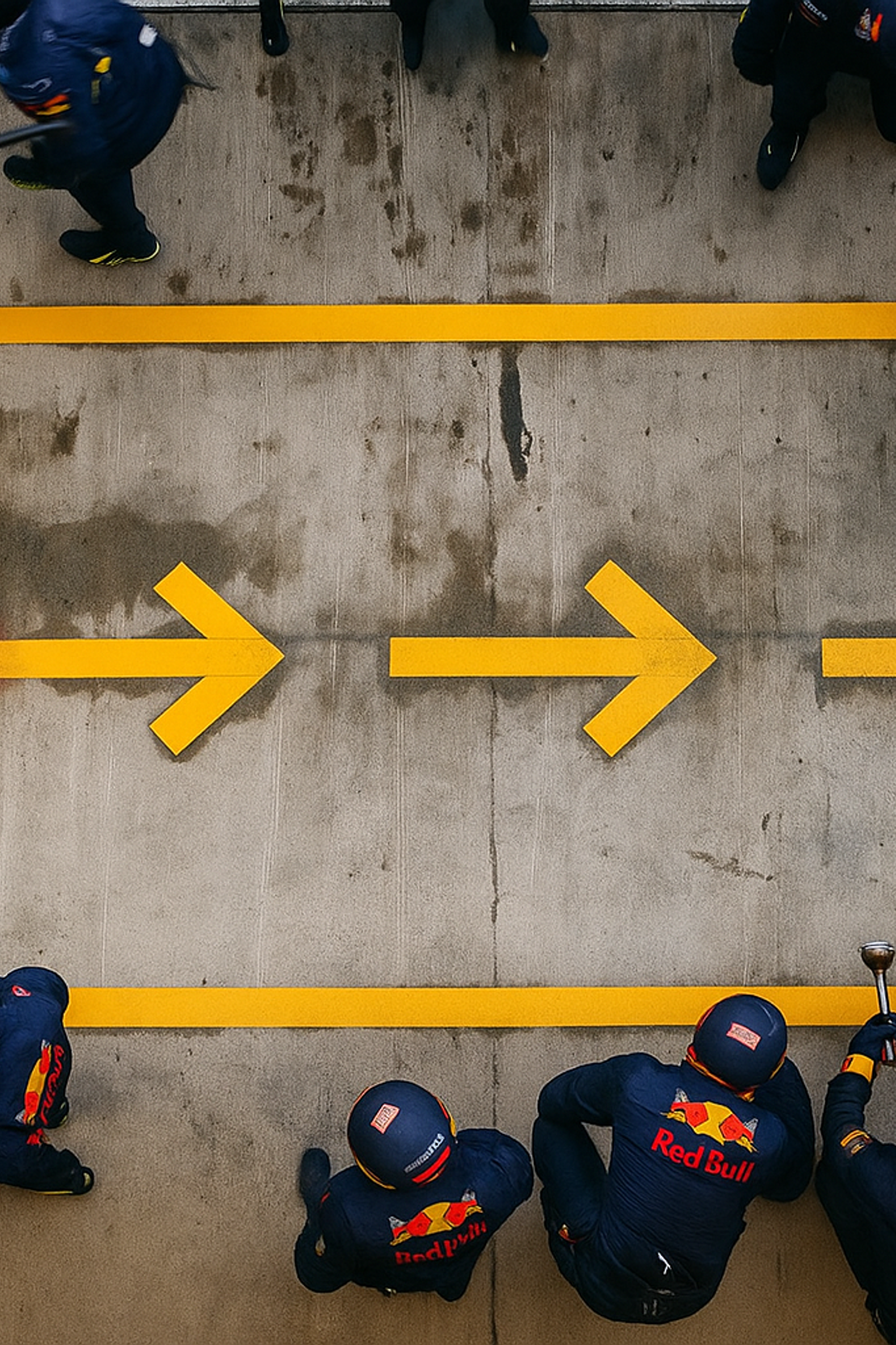 Draufsicht auf ein unscharfes F1-Auto, das an einer Red Bull-Boxencrew auf einer Betonstrecke mit gelben Richtungspfeilen vorbeirast.