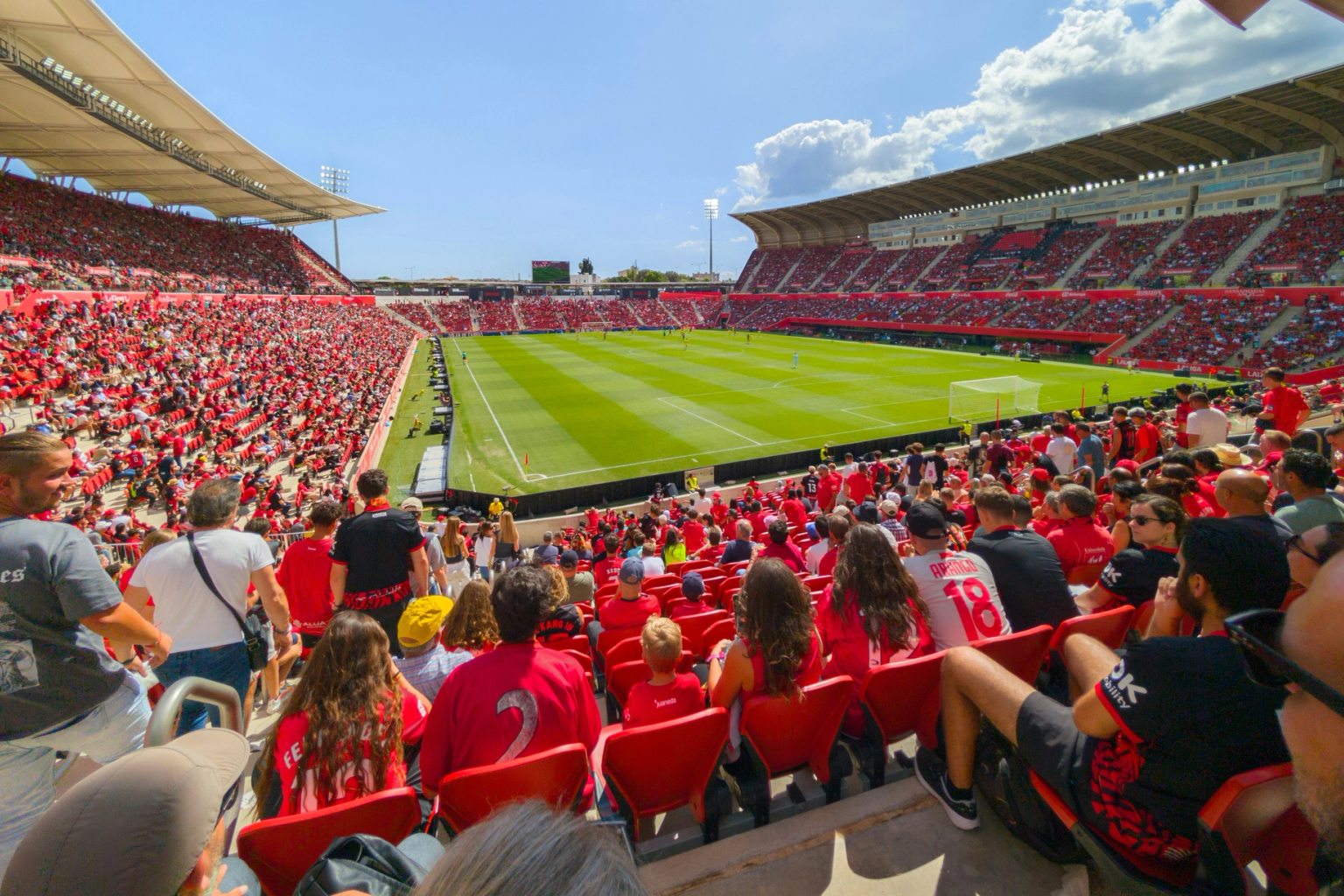 Un gran estadio lleno de personas vestidas de rojo viendo un partido de fútbol en un día soleado.