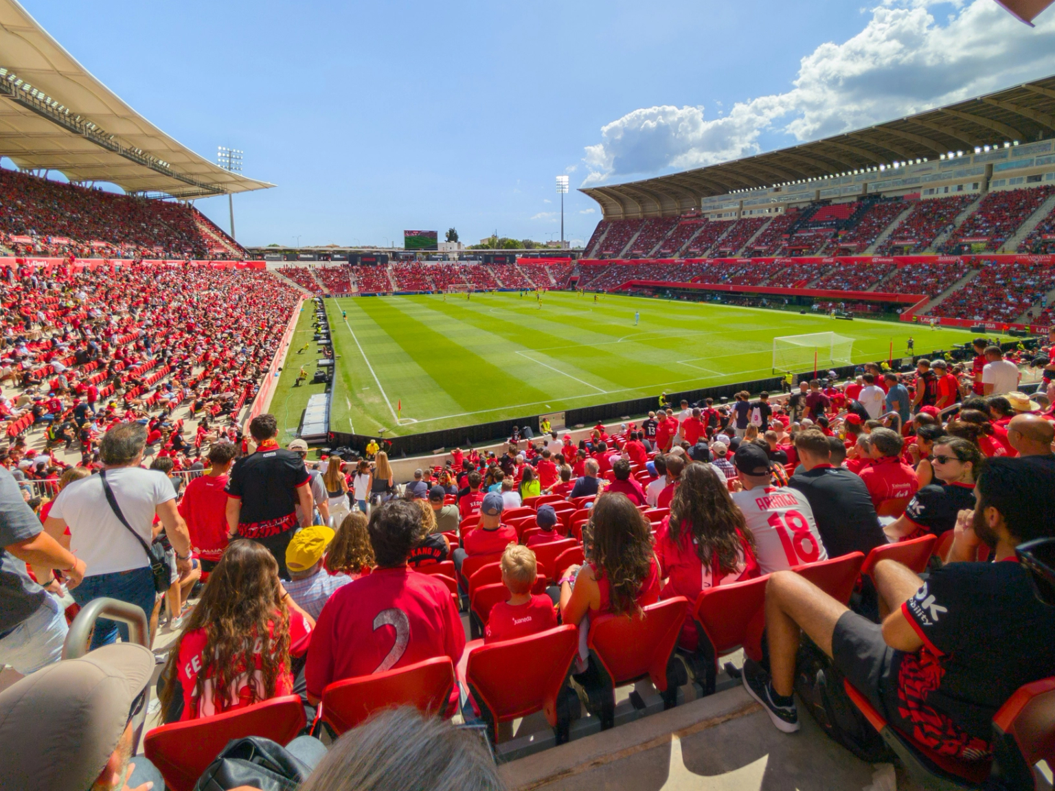 En stor stadion full med människor i rött som tittar på en fotbollsmatch på en solig dag.