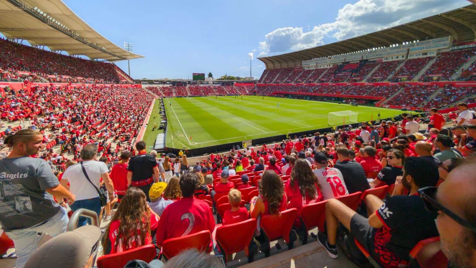 Un gran estadio lleno de personas vestidas de rojo viendo un partido de fútbol en un día soleado.