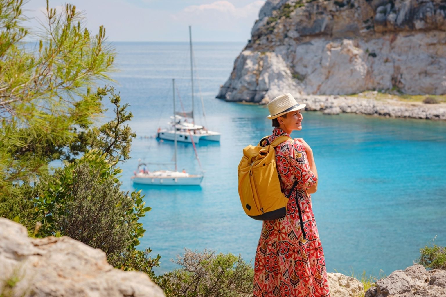 Kvinna med hatt och ryggsäck som blickar ut över en klarblå vik med segelbåtar och klippiga klippor. Anthony Quinn Bay, Rhodos