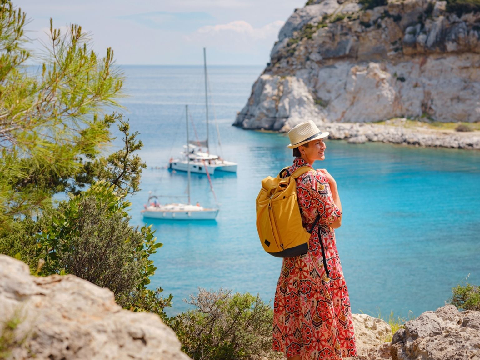 Kvinde med hat og rygsæk, der ser ud over en klar blå bugt med sejlbåde og klippefyldte klipper. Anthony Quinn Bay, Rhodos