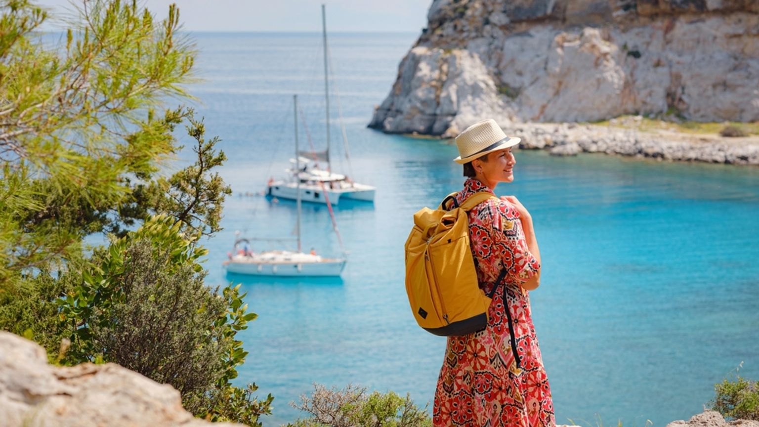Kvinna med hatt och ryggsäck som blickar ut över en klarblå vik med segelbåtar och klippiga klippor. Anthony Quinn Bay, Rhodos