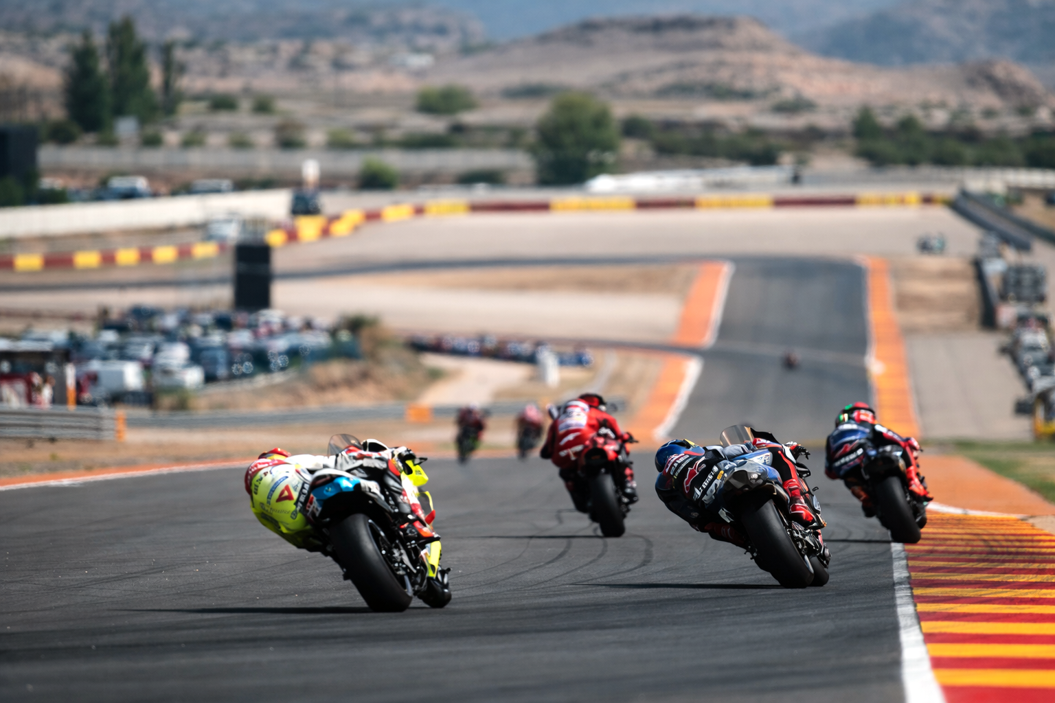 Motorcyklar lutar kraftigt in i en kurva på en solbelyst racerbana, med avlägsna kullar i bakgrunden.