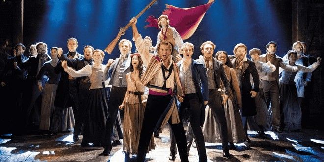 A dramatic stage performance featuring a large cast in period costumes, with a man holding a rifle and a woman waving a red flag.