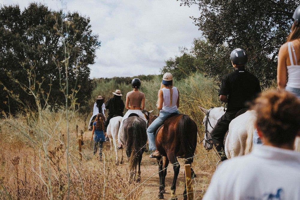 En grupp människor rider på hästar på en grusväg genom ett fält med högt buskage.