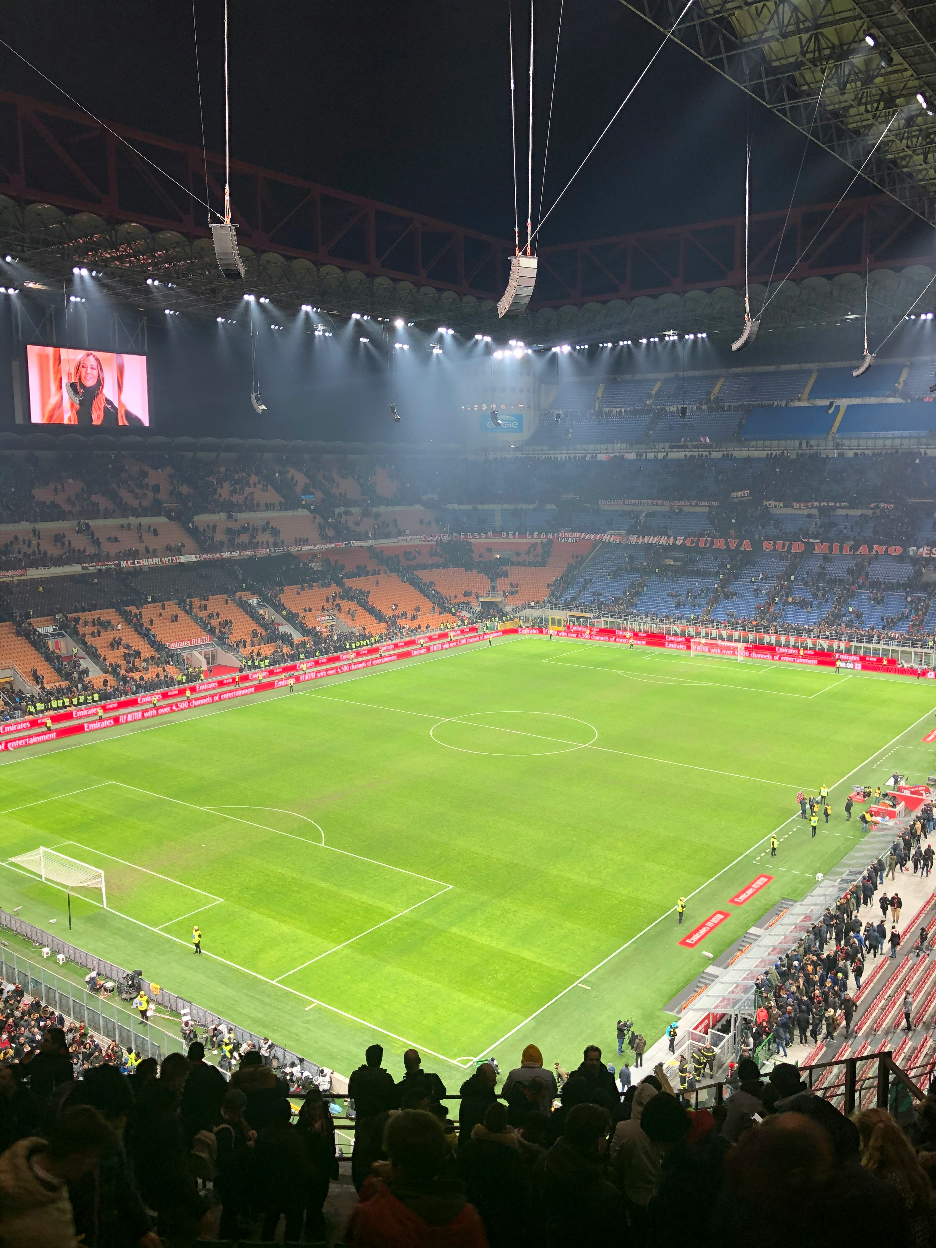 A brightly lit soccer stadium at night, with a green field, full stands, and a large screen showing a woman.