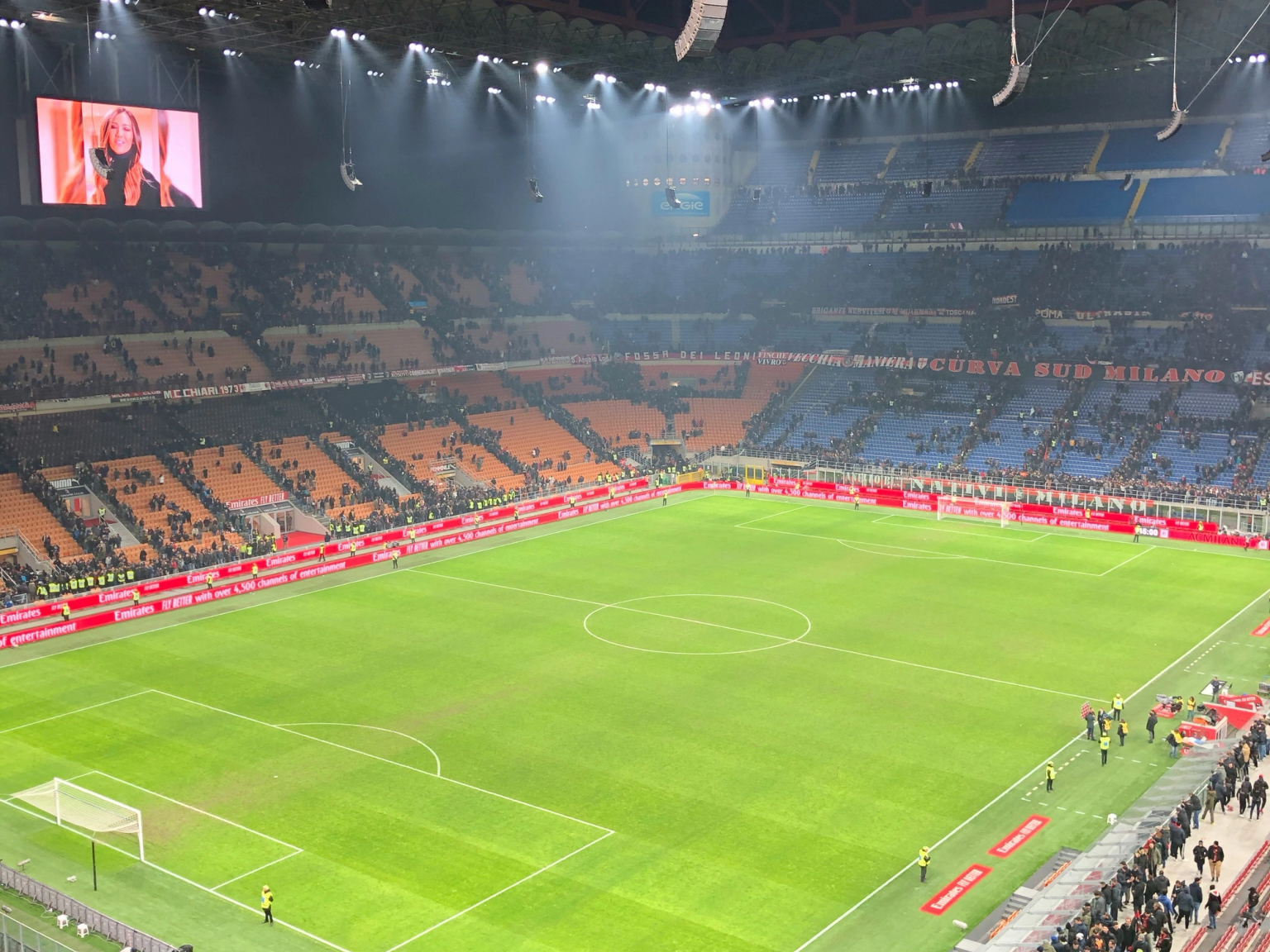 A brightly lit soccer stadium at night, with a green field, full stands, and a large screen showing a woman.