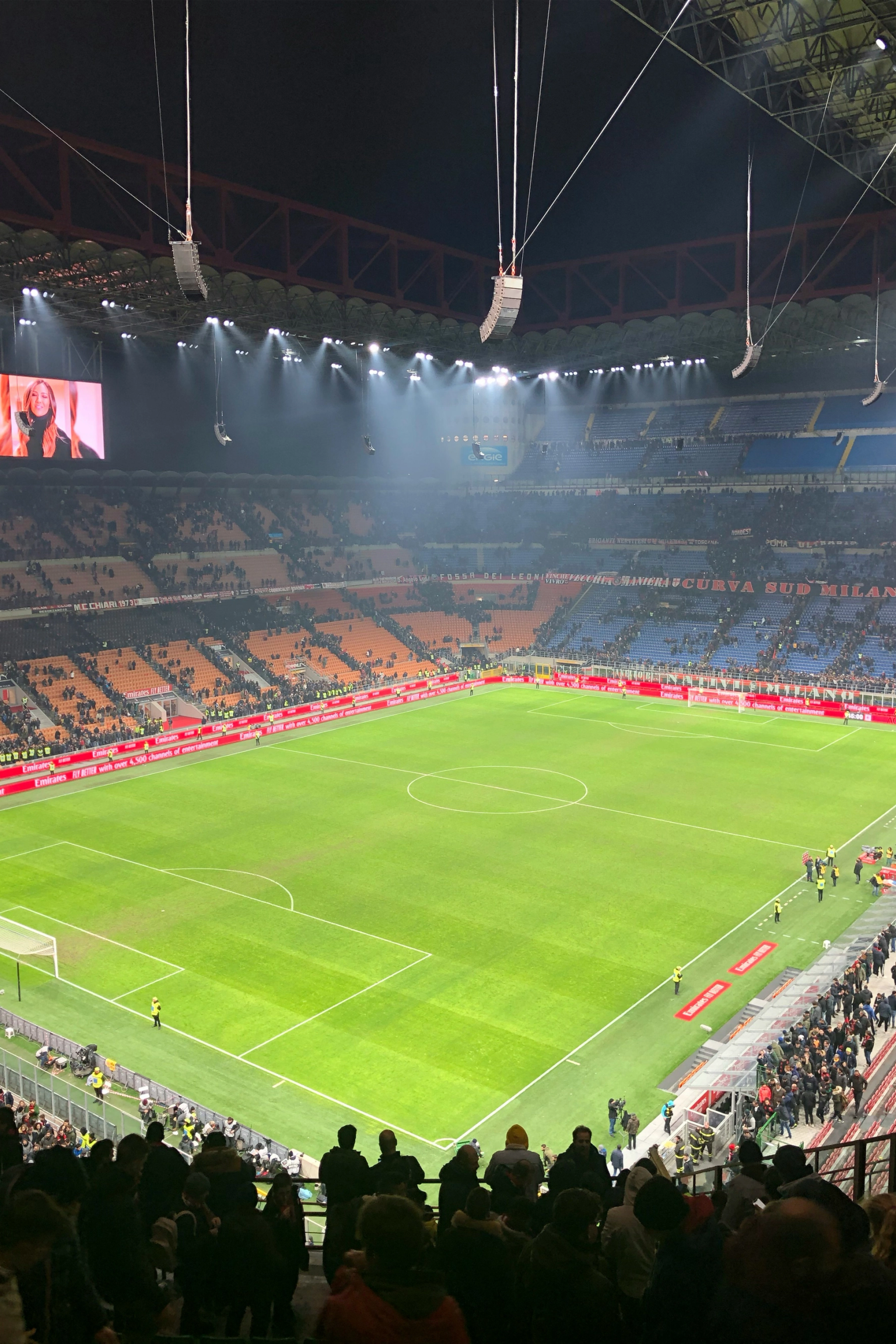 A brightly lit soccer stadium at night, with a green field, full stands, and a large screen showing a woman.
