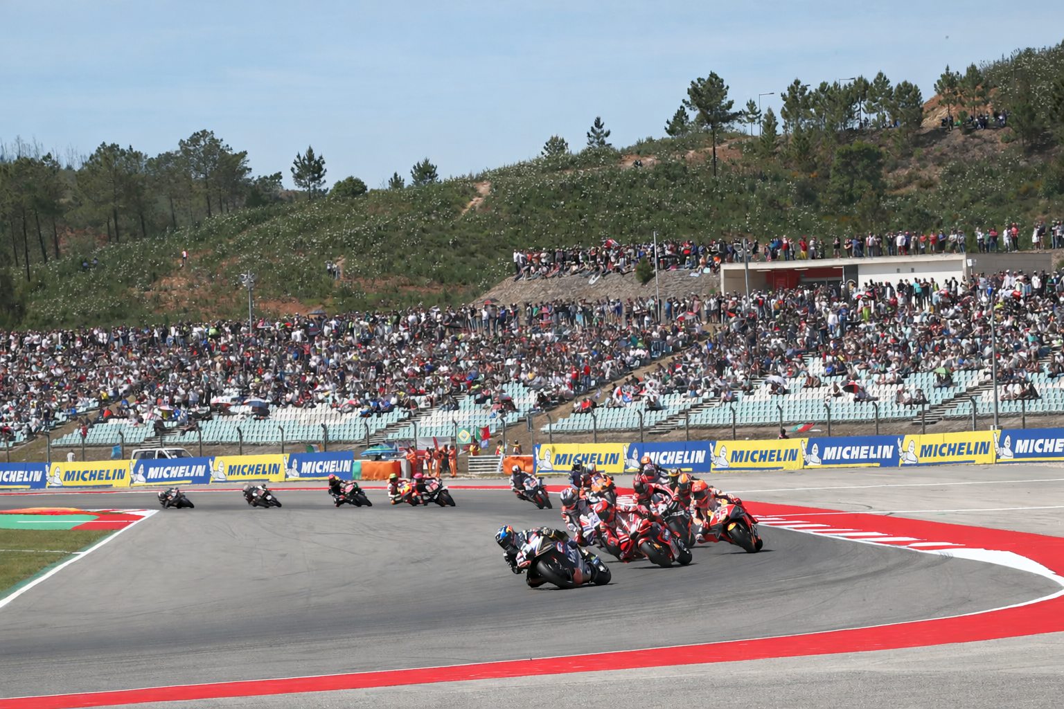 Motocicletas correm em alta velocidade numa curva da pista, observadas por uma grande multidão nas arquibancadas.
