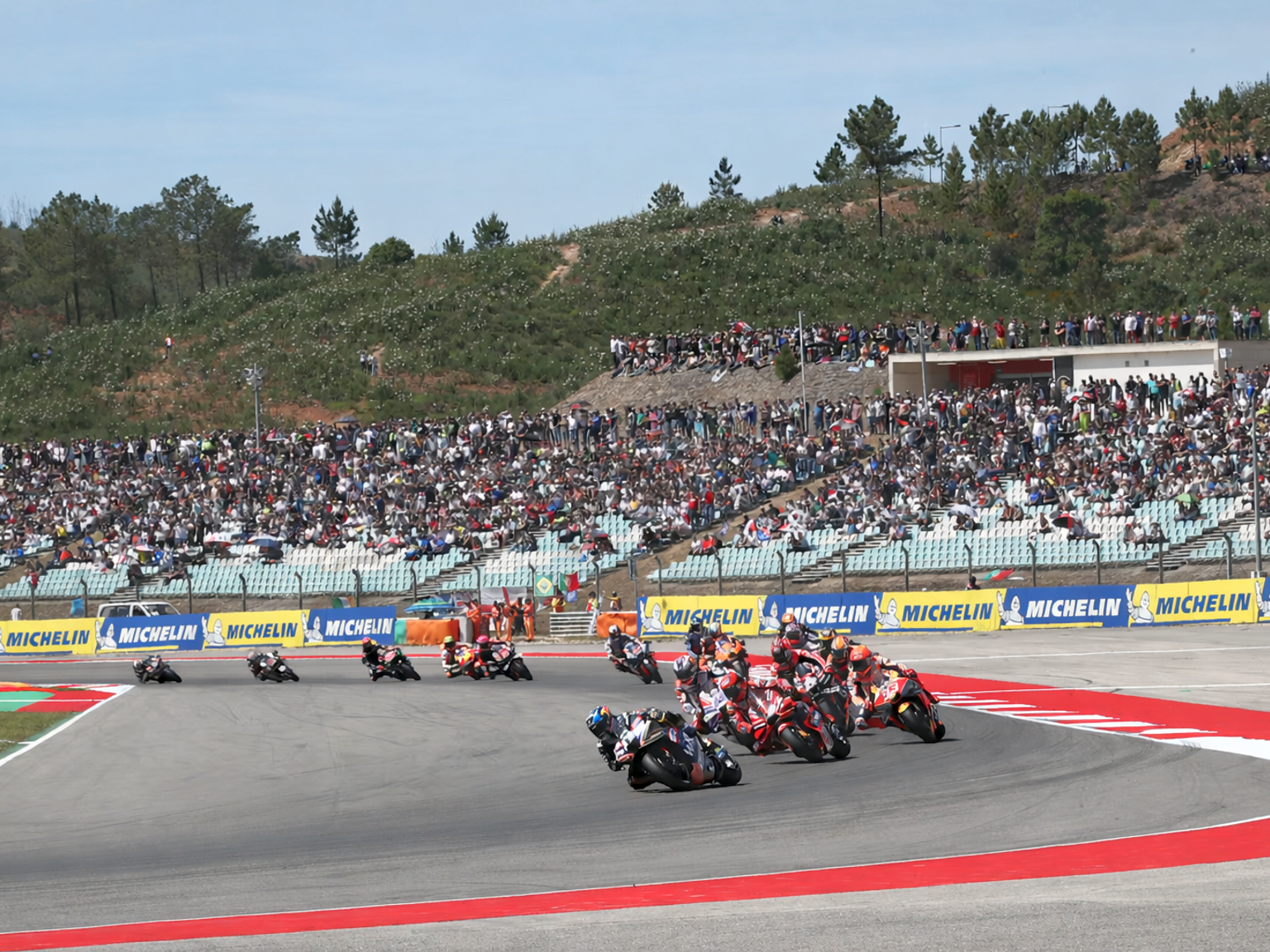 Motocicletas correm em alta velocidade numa curva da pista, observadas por uma grande multidão nas arquibancadas.
