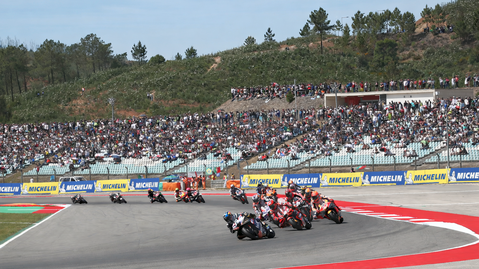Varias motocicletas corren alrededor de una curva en una pista, observadas por una gran multitud en las gradas.