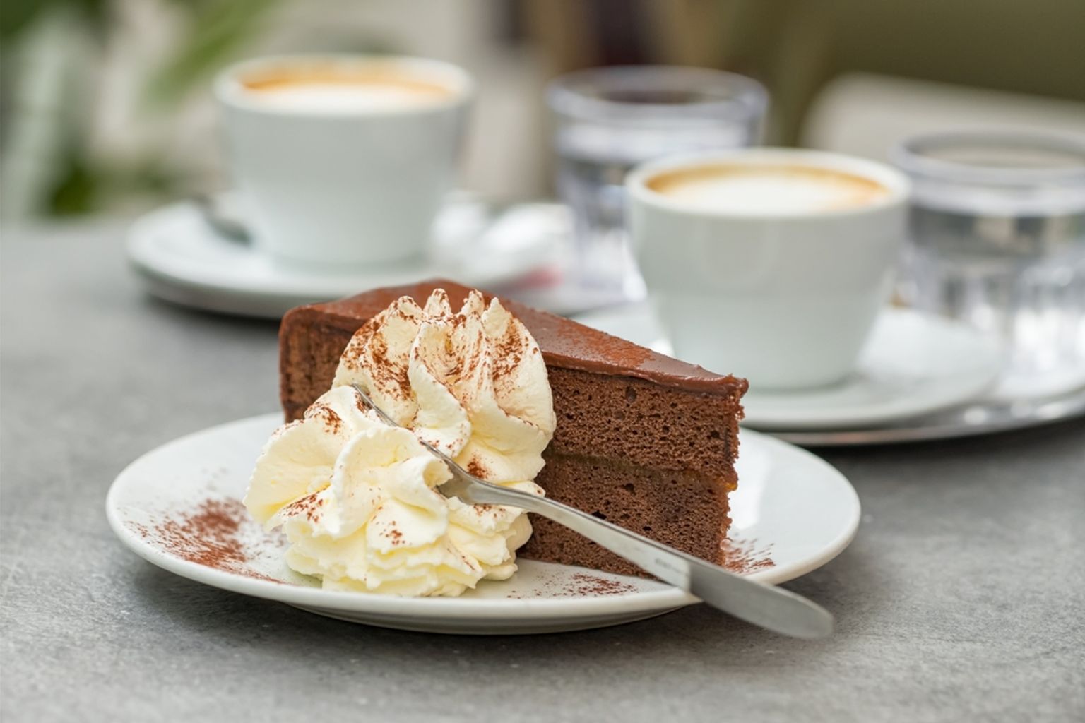 En skiva chokladkaka med vispgrädde på en tallrik, med kaffe och vattenglas i bakgrunden.
