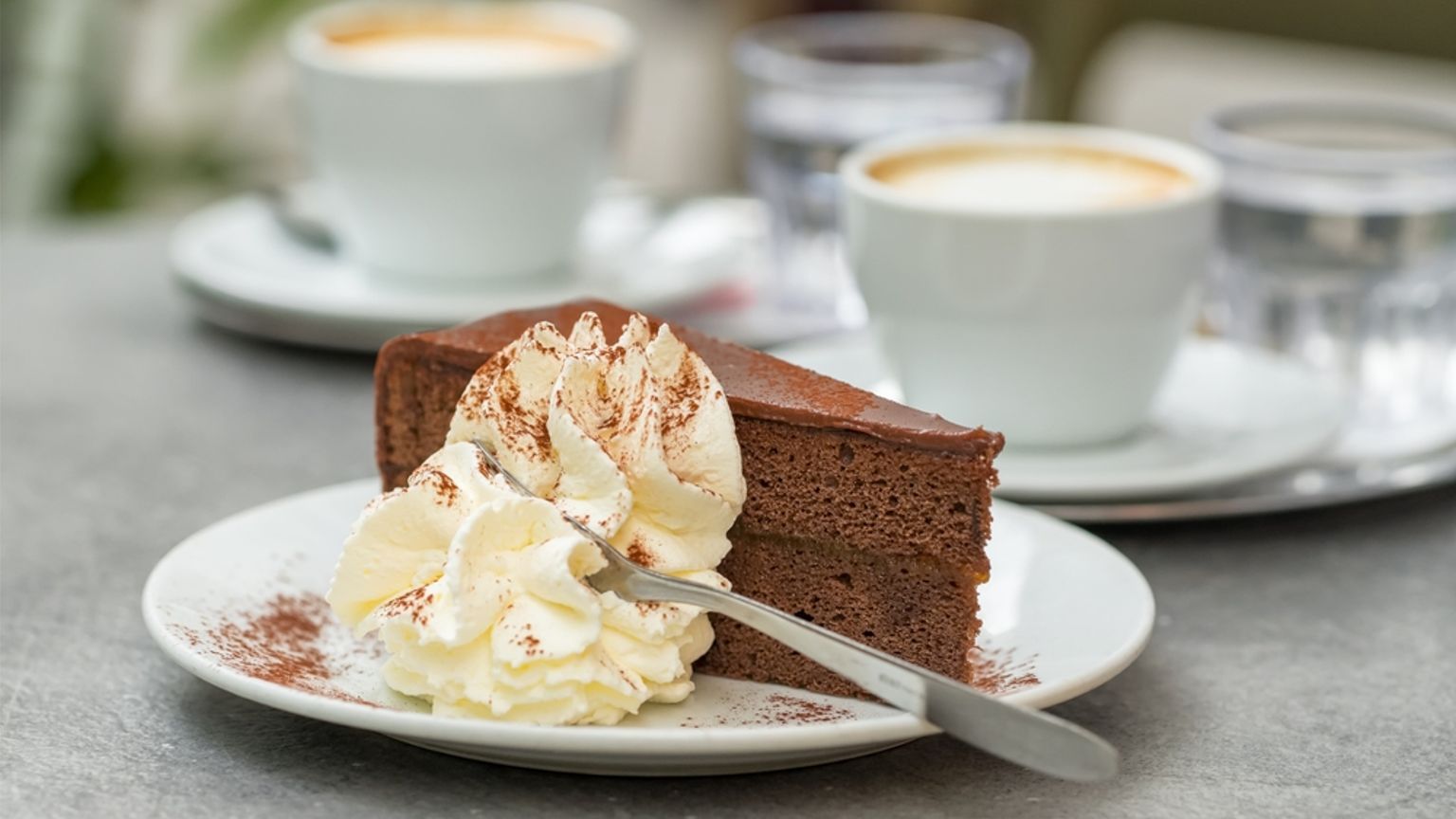 En skiva chokladkaka med vispgrädde på en tallrik, med kaffe och vattenglas i bakgrunden.