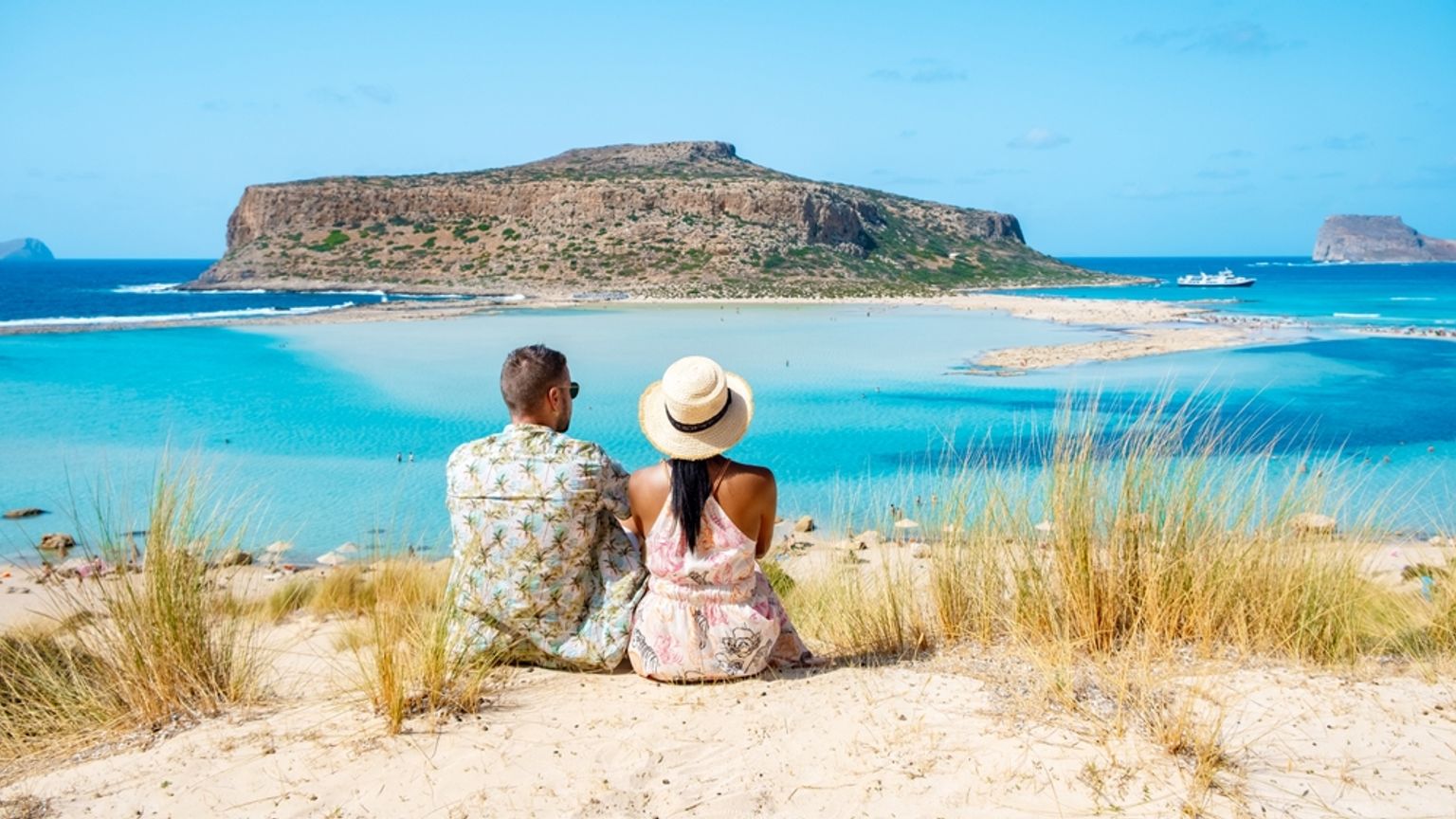 A couple sits on a sand dune, admiring a vibrant turquoise lagoon and a large rocky island.