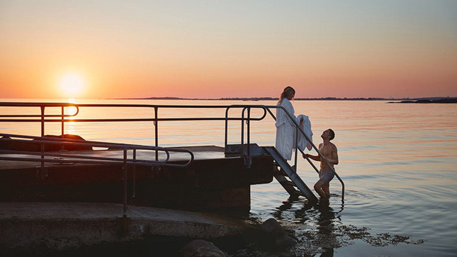 en man och en kvinna står på en brygga i vattnet vid solnedgången