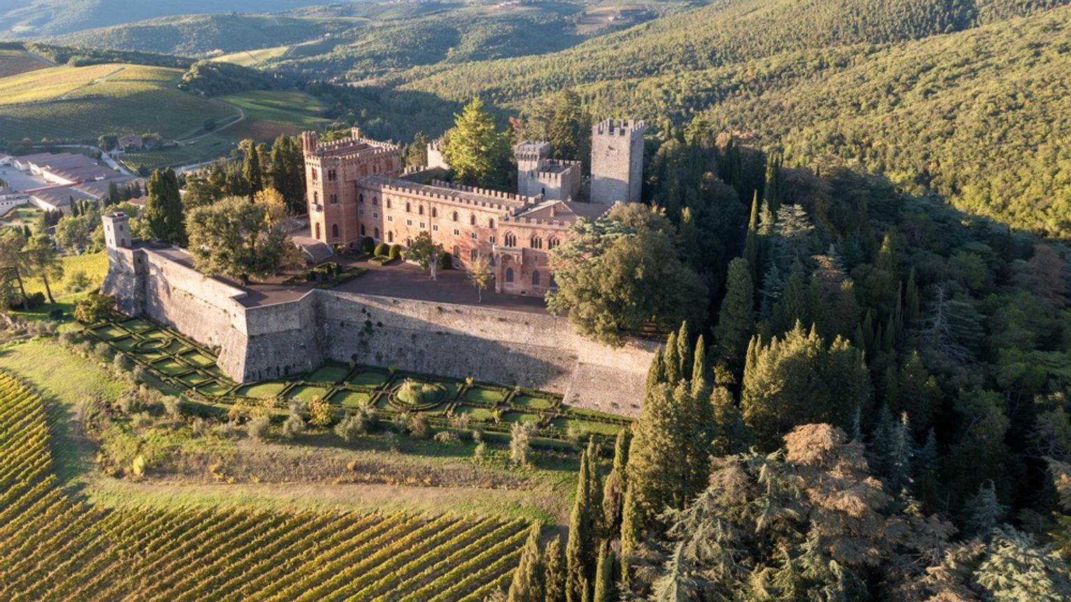 Luftfoto af et stenfort og en lang mur, der ligger omgivet af vinmarker og grønne, skovklædte bakker. Chianti, Italien