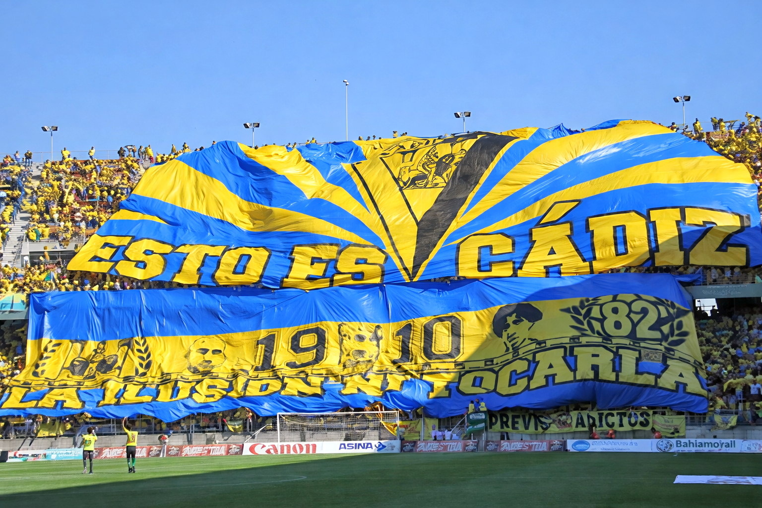 Duże niebiesko-żółte tifo z napisem „ESTO ES CÁDIZ” rozwinięte nad tłumem kibiców piłkarskich na stadionie.