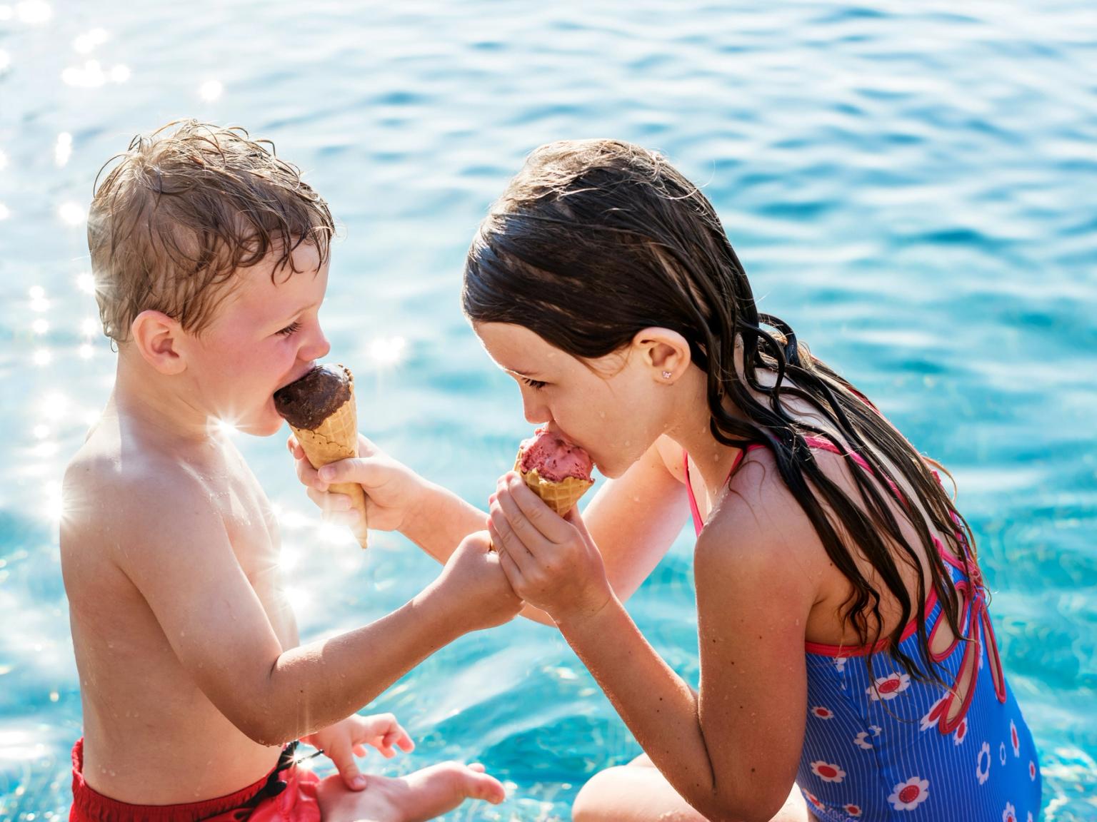 En pojke och en flicka som äter glasstrutar vid gnistrande blått vatten.