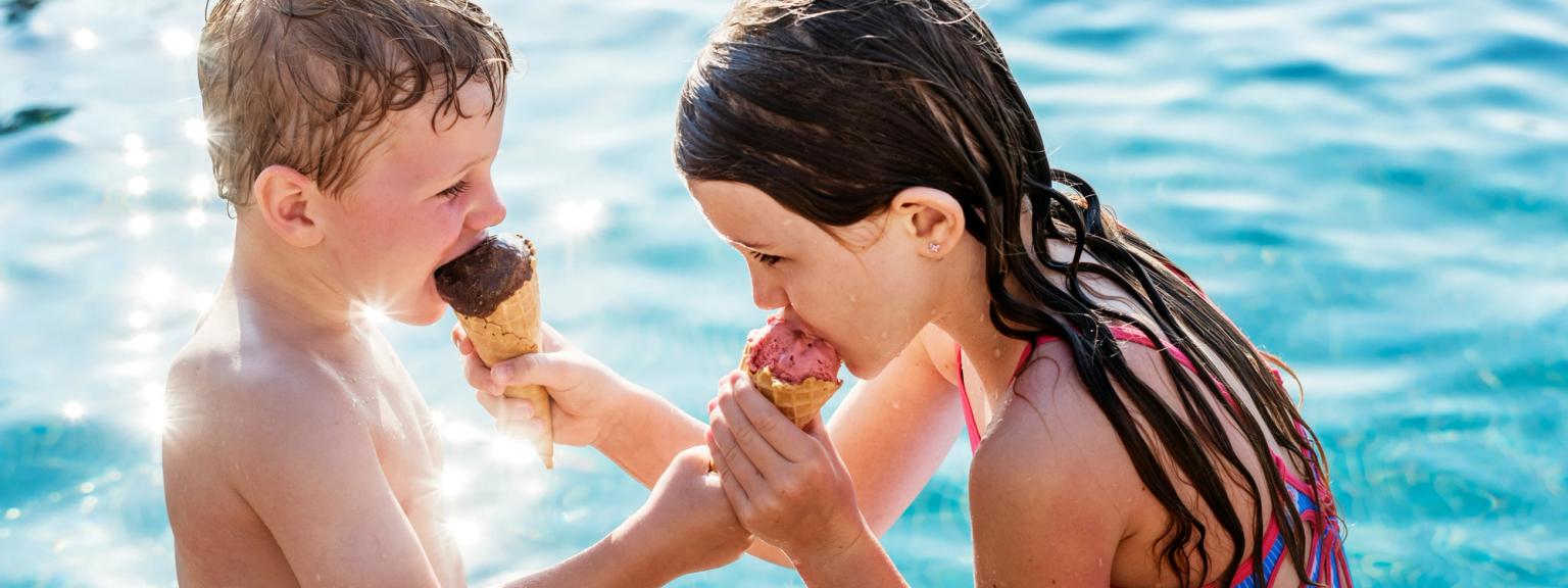 En pojke och en flicka som äter glasstrutar vid gnistrande blått vatten.