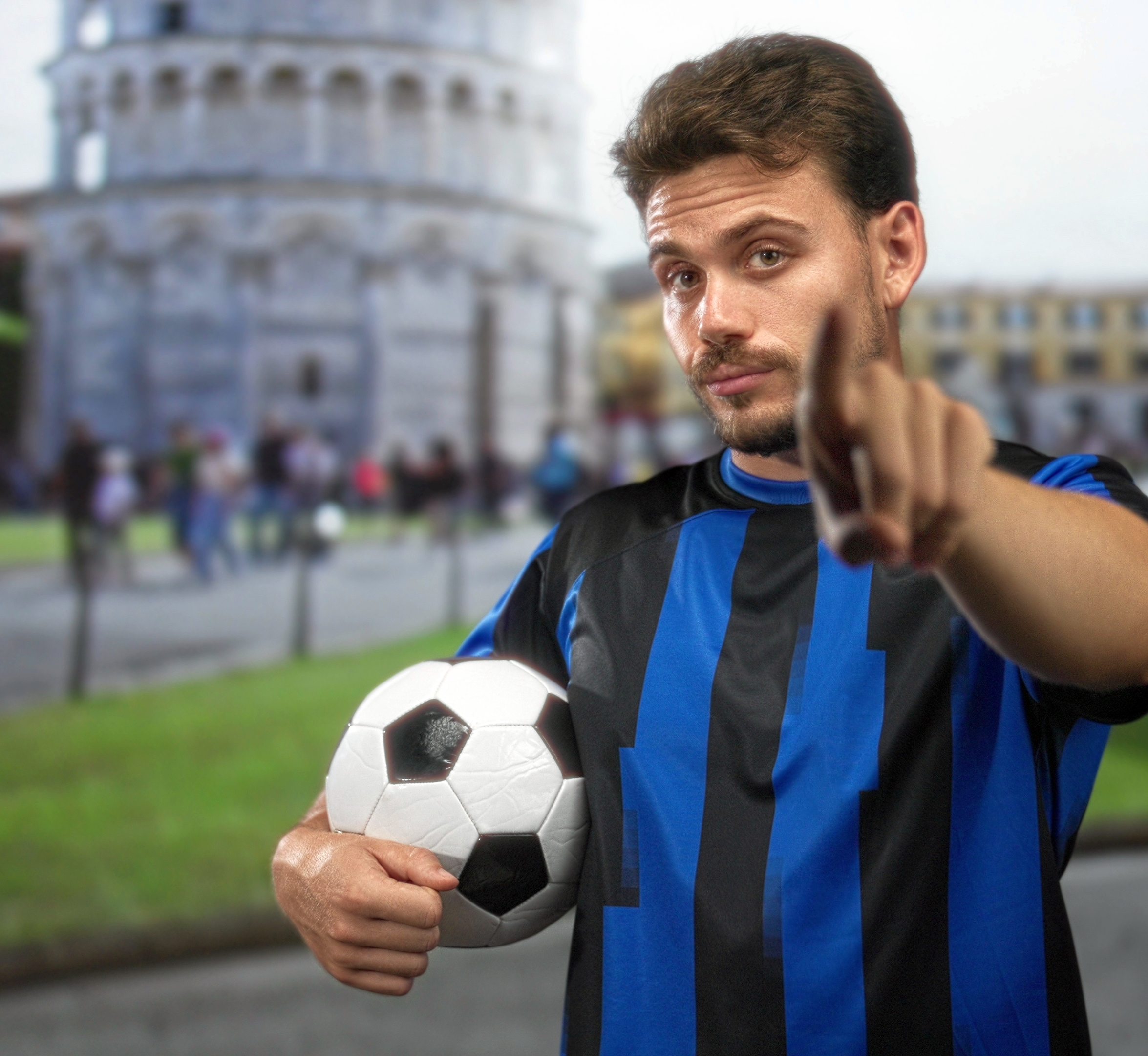 Man in blue and black soccer jersey holds a ball and points at the viewer, with the Leaning Tower of Pisa in the blurry background.