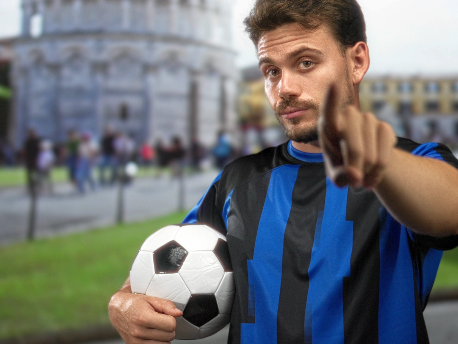 Man in blue and black soccer jersey holds a ball and points at the viewer, with the Leaning Tower of Pisa in the blurry background.