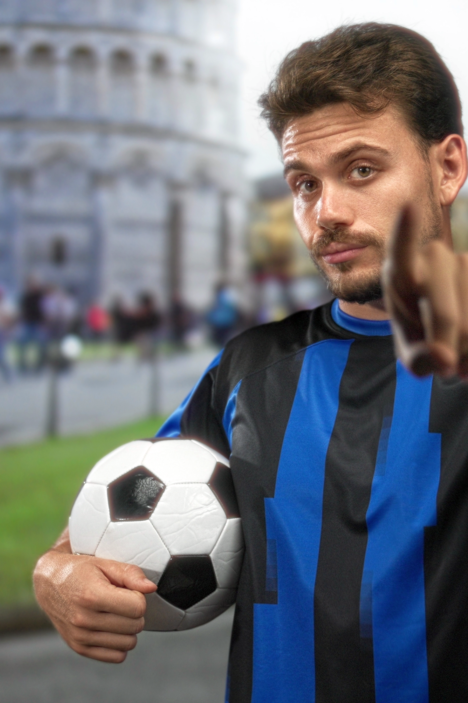 Man in blue and black soccer jersey holds a ball and points at the viewer, with the Leaning Tower of Pisa in the blurry background.