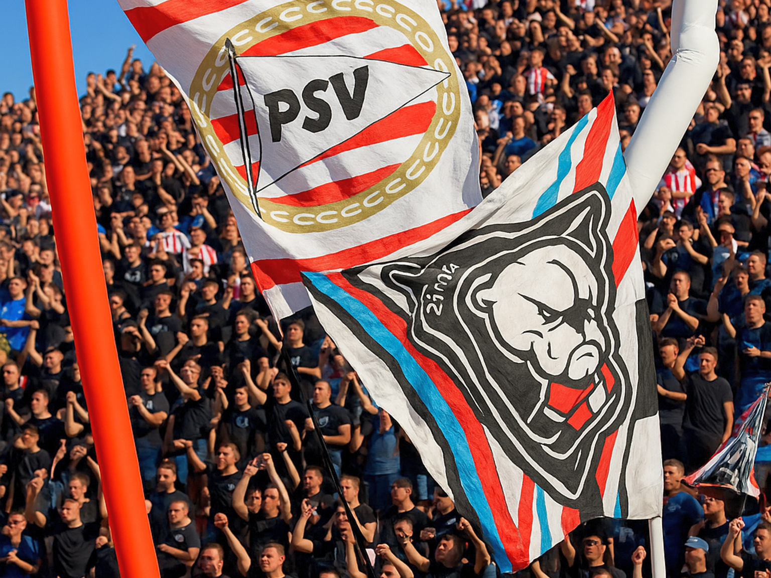 Två stora flaggor, en med PSV-märke och en annan med en bulldoggdesign, vajar ovanför en stadionpublik med höjda nävar.