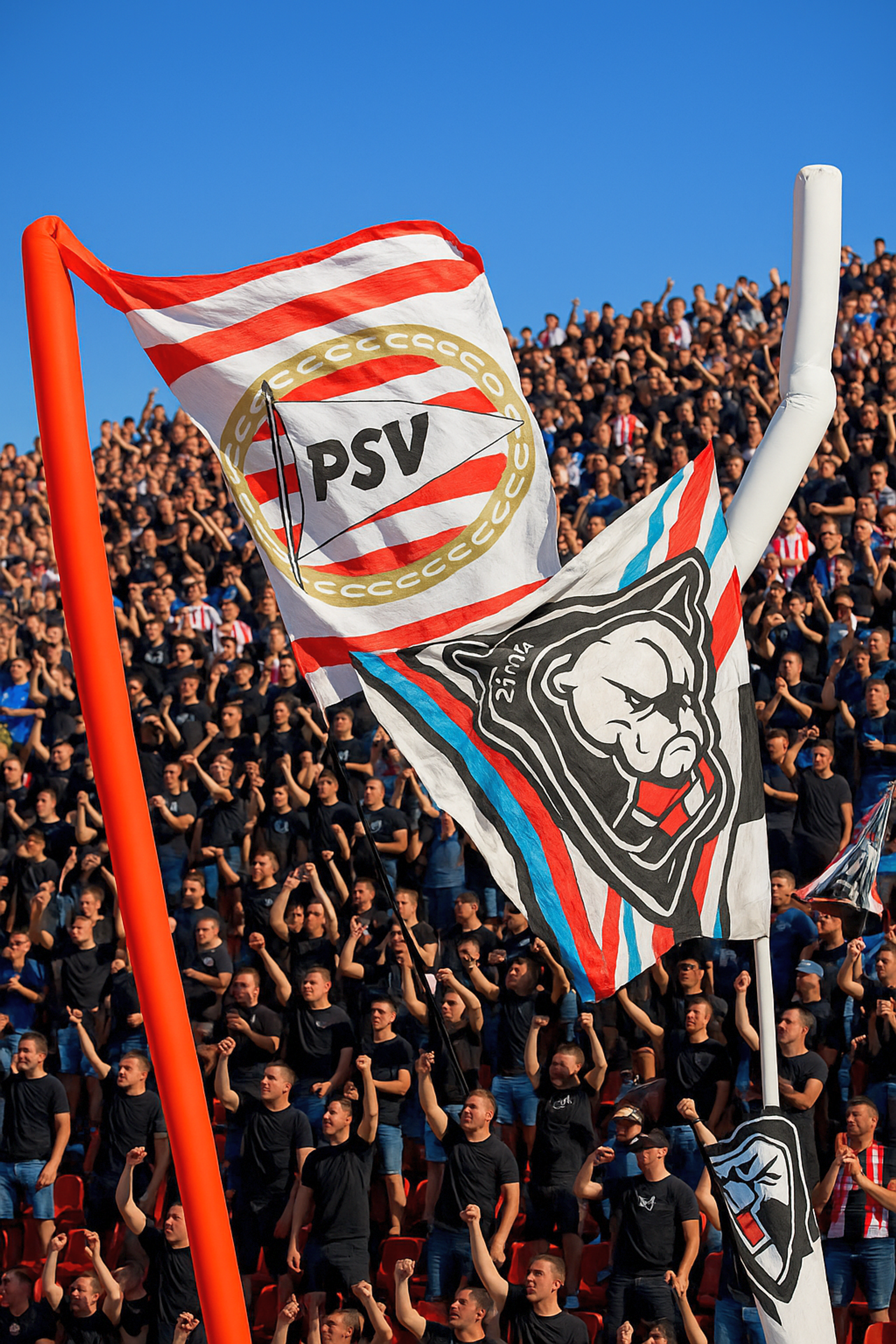 Två stora flaggor, en med PSV-märke och en annan med en bulldoggdesign, vajar ovanför en stadionpublik med höjda nävar.