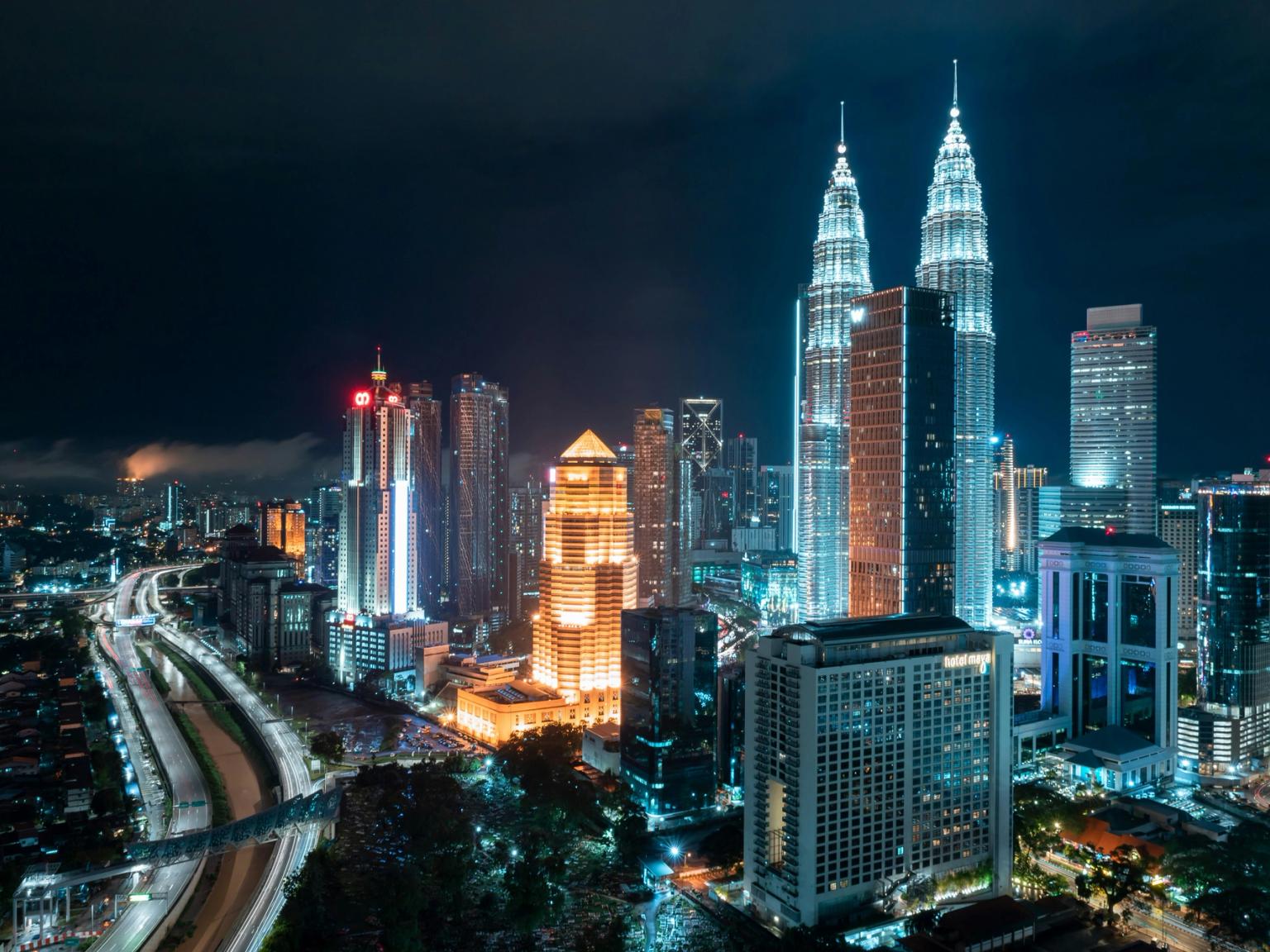 Kuala Lumpurs bybillede om natten med oplyste Petronas Twin Towers, omkringliggende skyskrabere og en motorvej, der viser lysstriber.