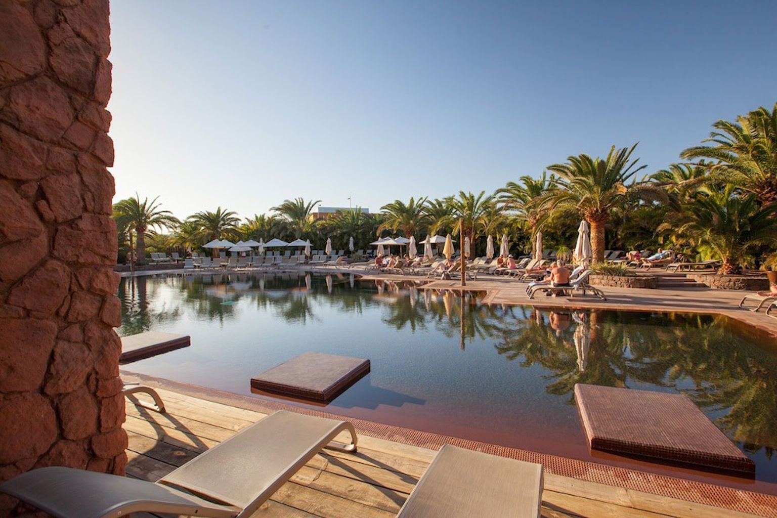 En stor resort swimmingpool med mørkt vand, flydende platforme, palmer og solbadere.