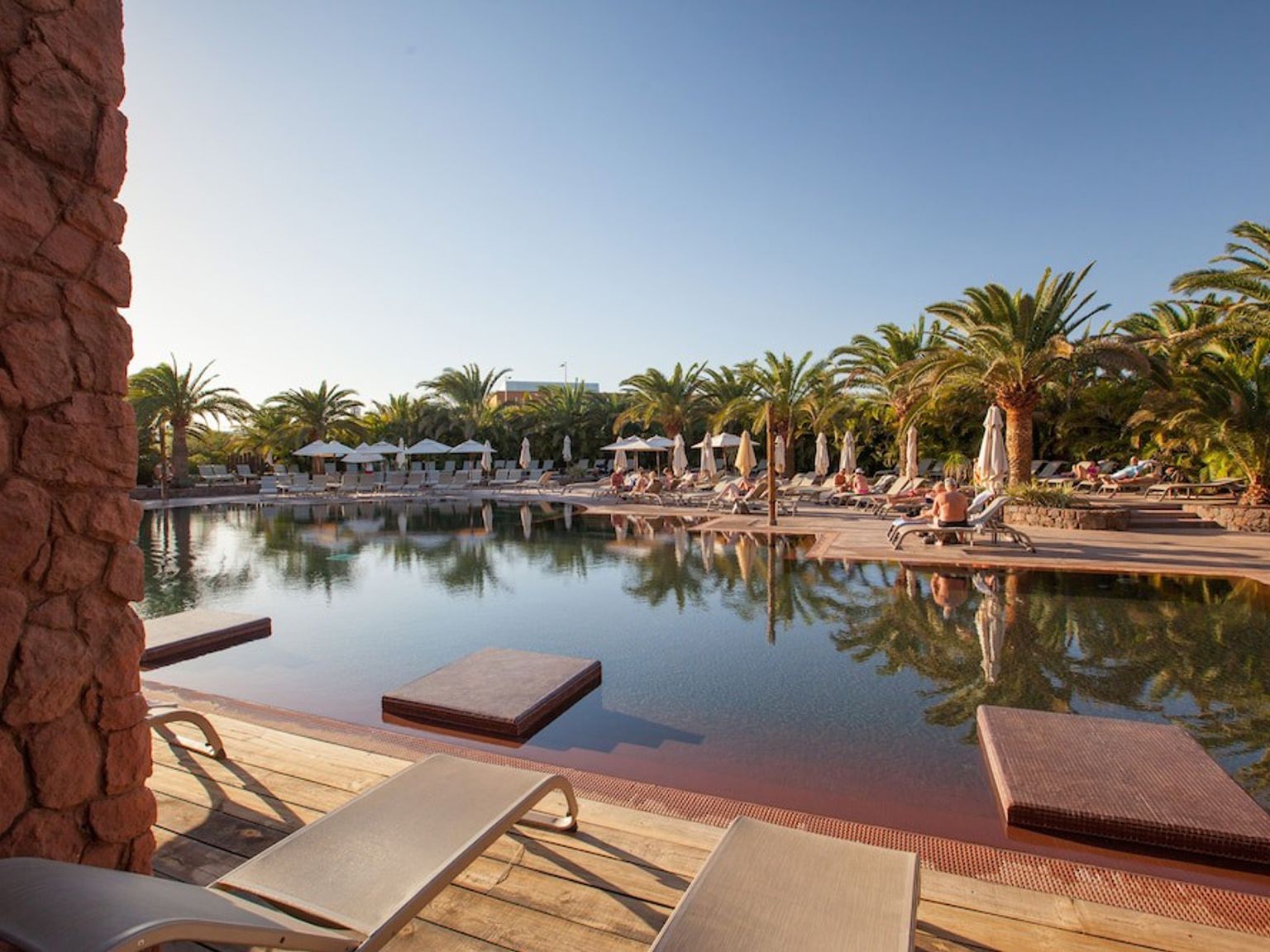 En stor resort swimmingpool med mørkt vand, flydende platforme, palmer og solbadere.