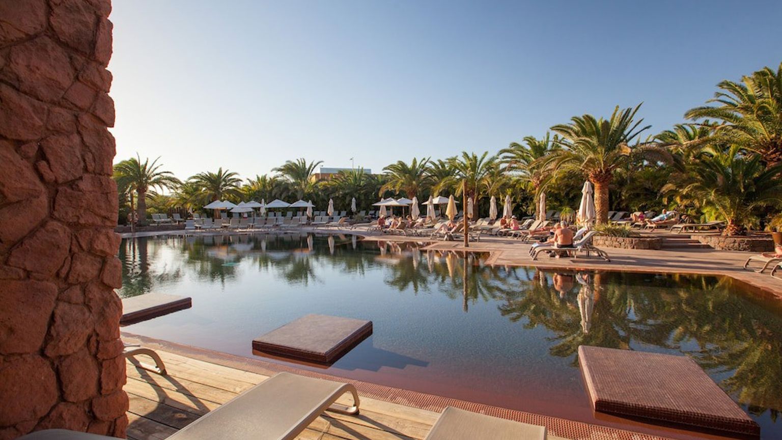 En stor resort swimmingpool med mørkt vand, flydende platforme, palmer og solbadere.