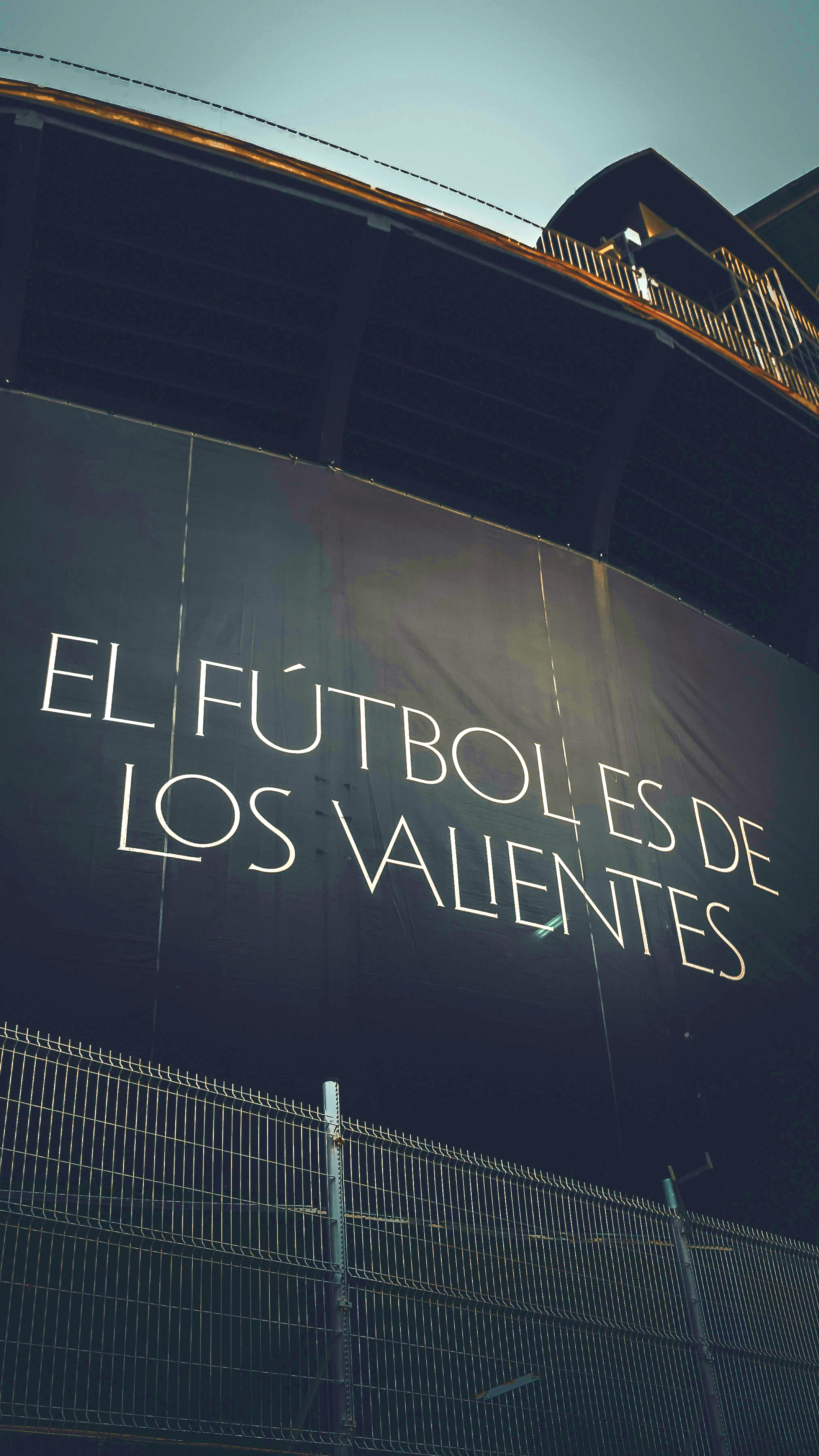 A black banner on a stadium reads "EL FÚTBOL ES DE LOS VALIENTES" (Football is for the brave) in white text, with a metal fence below.