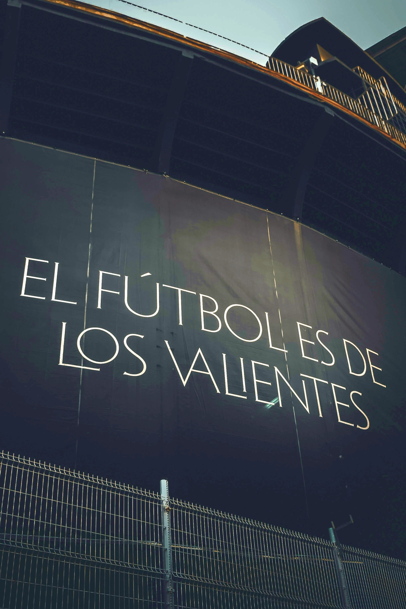 Et sort banner på et stadion lyder "EL FÚTBOL ES DE LOS VALIENTES" (Fodbold er for de modige) i hvid tekst, med et metalhegn nedenunder.
